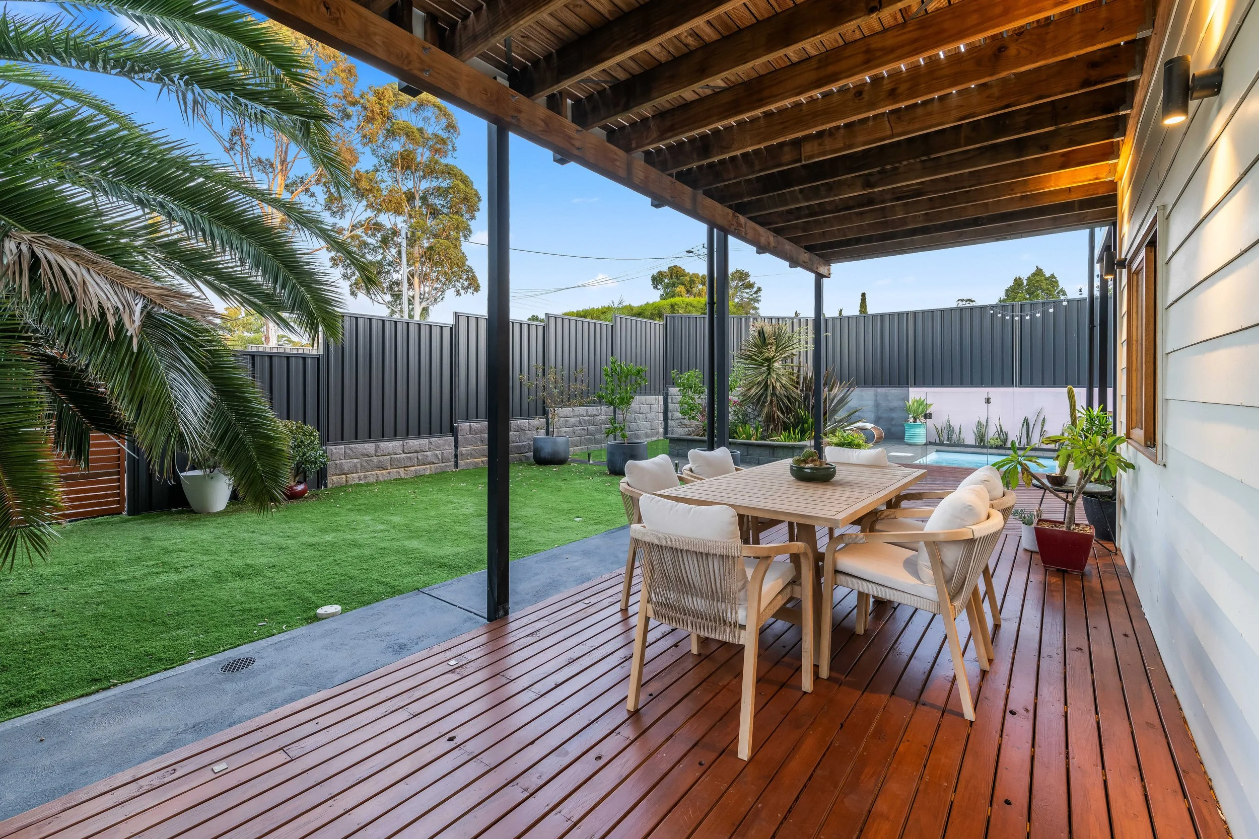 Covered outdoor patio with wooden floor and ceiling, rectangular dining table with chairs, lush green lawn, potted plants, tall trees, and a dark metal fence in the background.