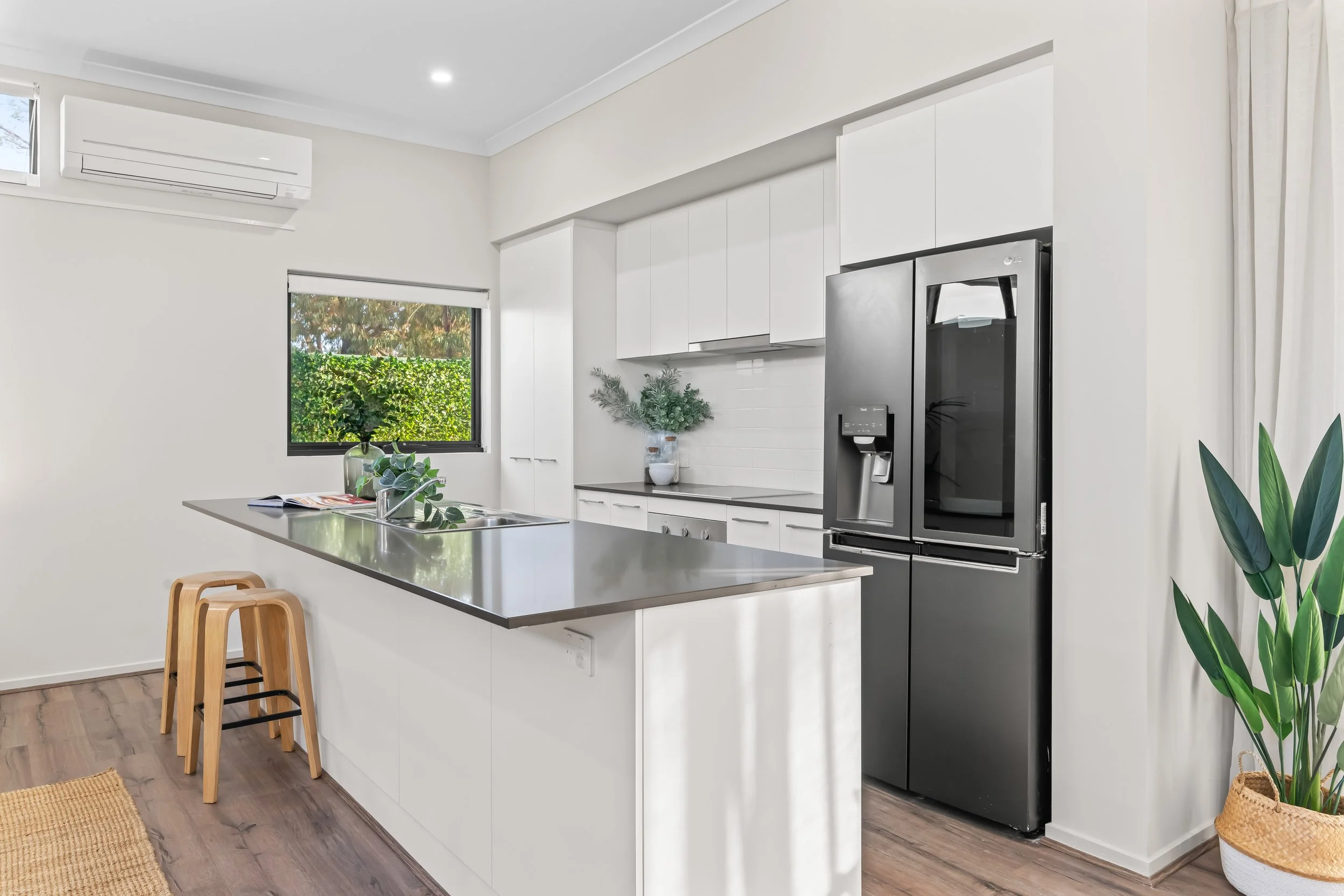 Modern kitchen with white cabinets, stainless steel refrigerator, green plants, window showing greenery outside, and wooden bar stools at a kitchen island.
