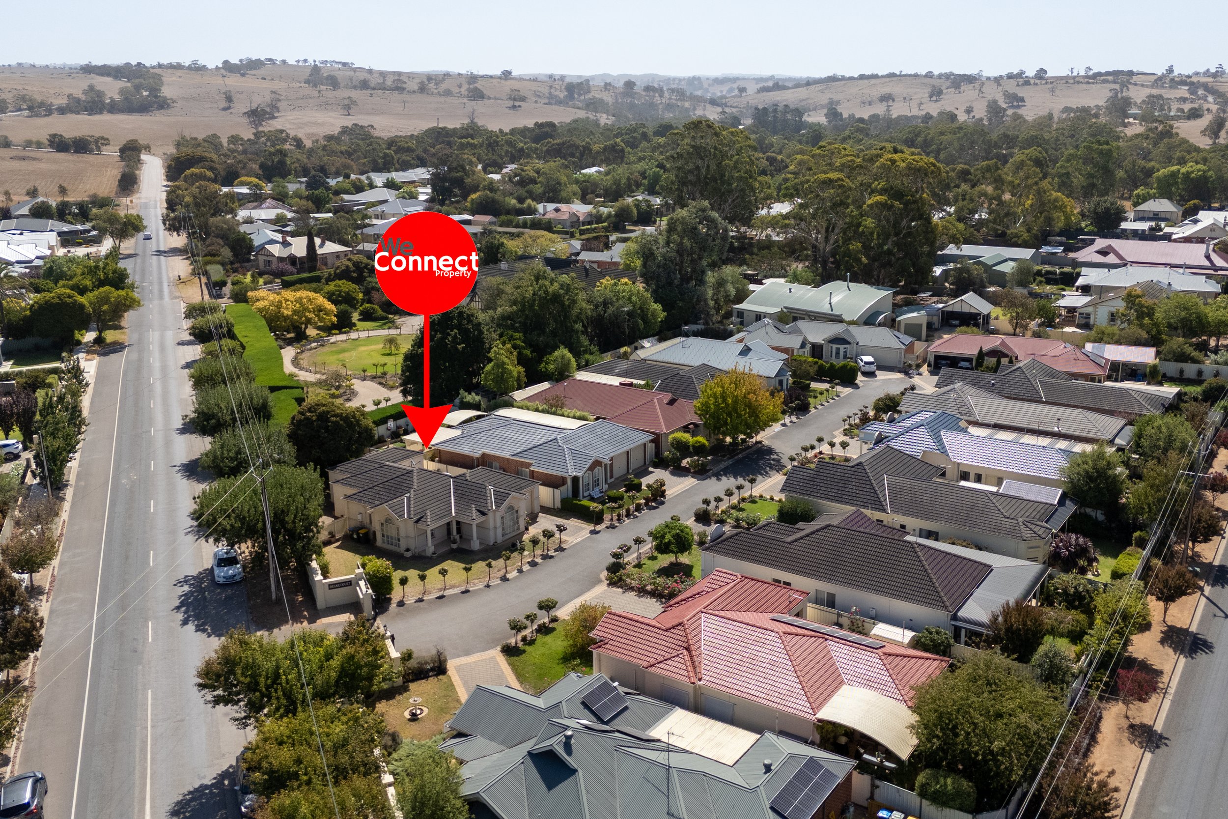 Aerial view of a suburban neighborhood with houses, trees, and a main road, with a hilly countryside in the background. A red 