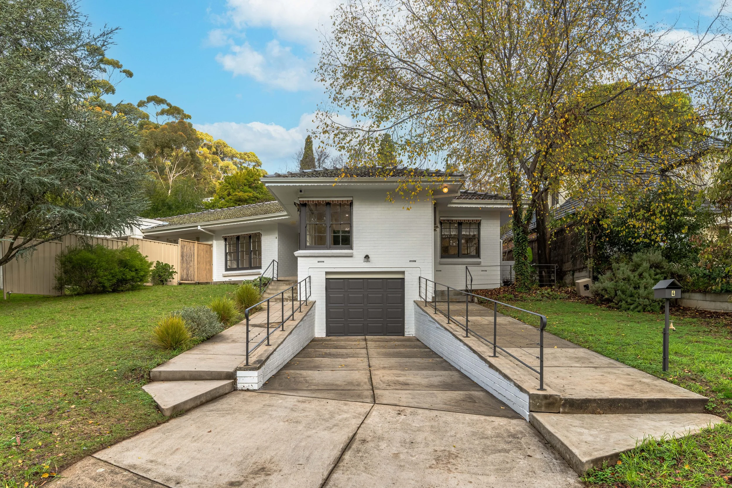 Front view of a suburban house with a staircase leading to the main entrance, surrounded by greenery and trees, under a partly cloudy sky.