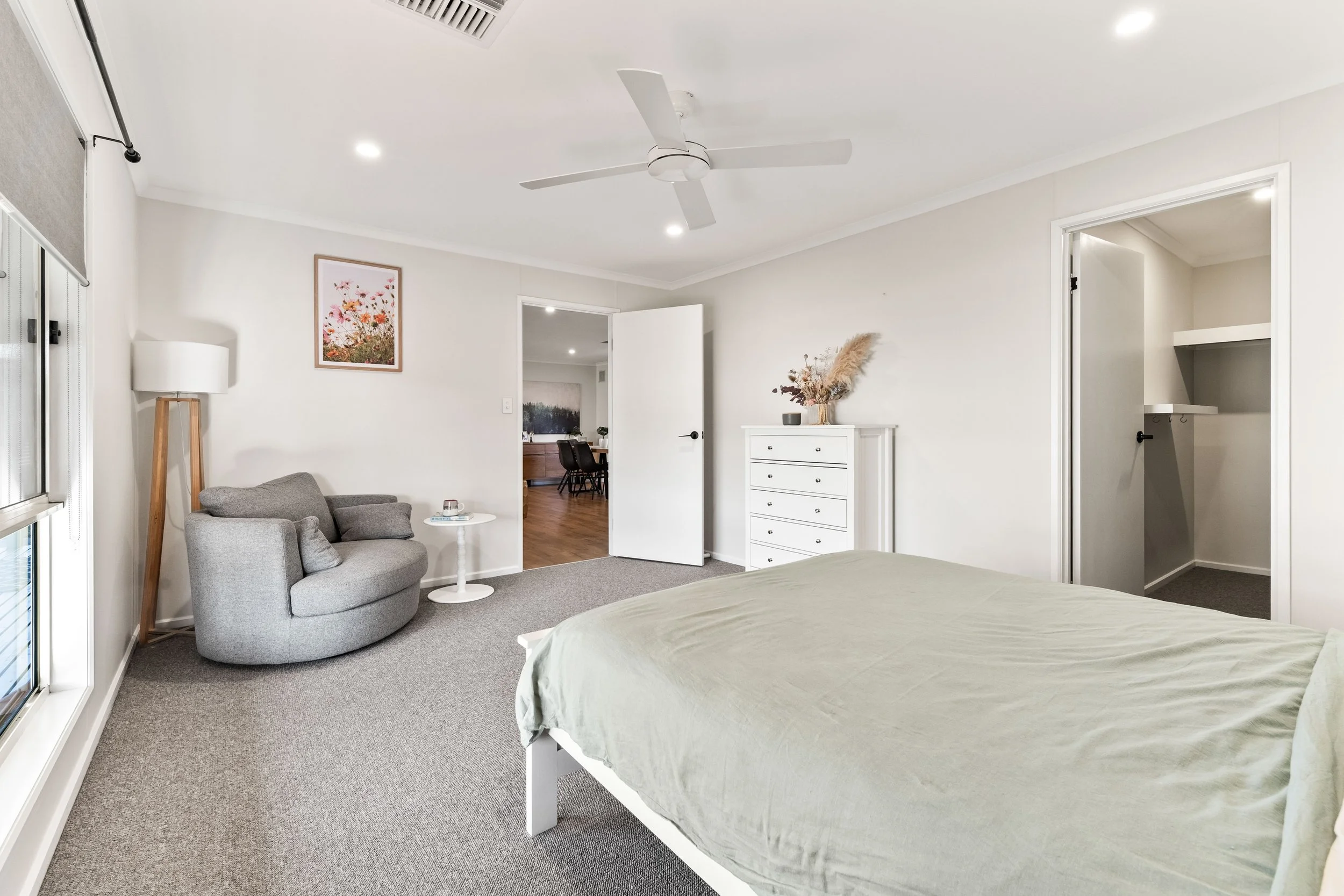 A bedroom with a bed in the foreground, a gray armchair, a small white round table, and a white dresser with decorative dried flowers. There is a white ceiling fan, a painting on the wall, a closet with open shelves, and an open door leading to a din