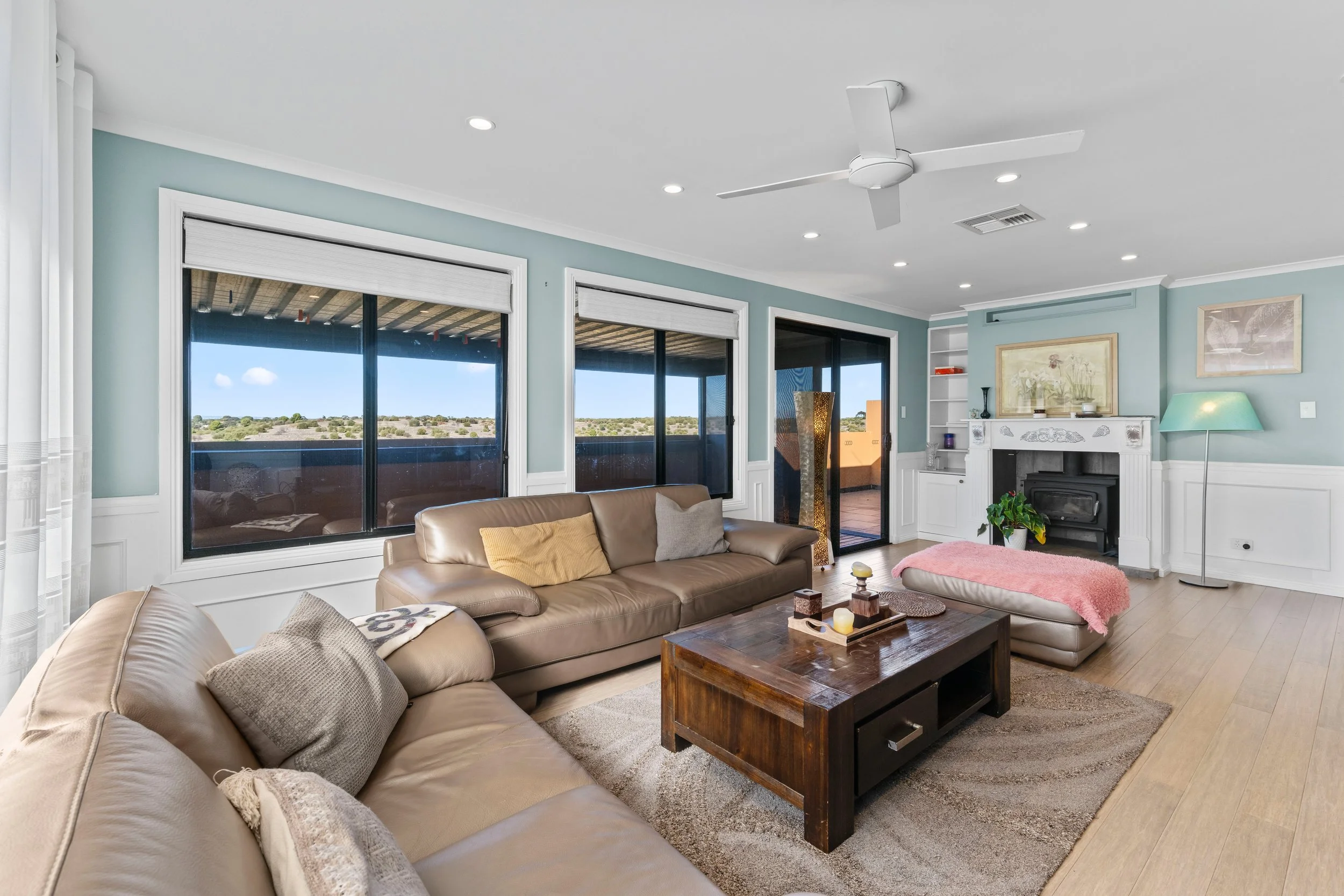 A living room with beige leather sofas, a wooden coffee table, large windows showing an outdoor landscape, a white fireplace with decorative items, a turquoise floor lamp, and artwork on the walls.