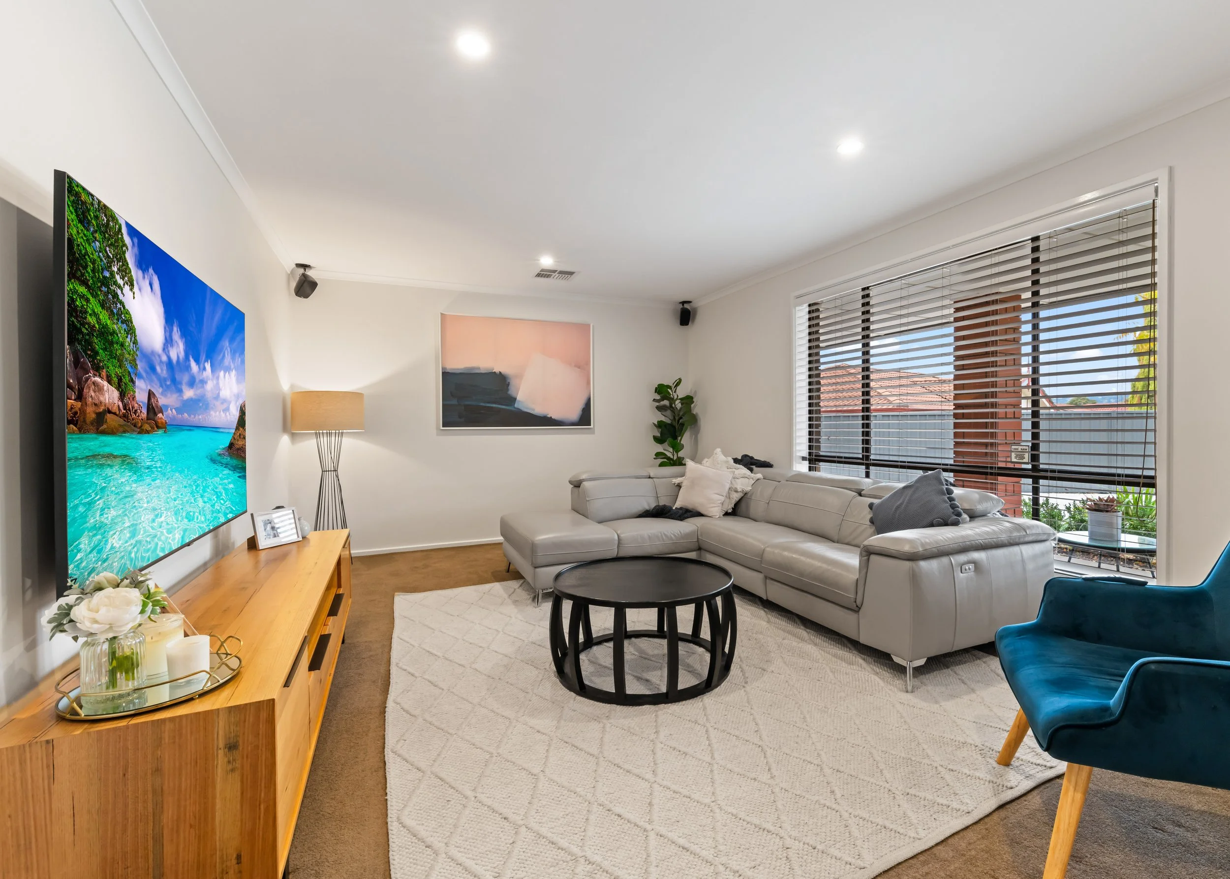 Living room with a gray sectional sofa, black round coffee table, blue armchair, TV displaying a tropical beach scene, modern artwork, tall floor lamp, plant, large window with blinds, and decorative items on a wooden console.