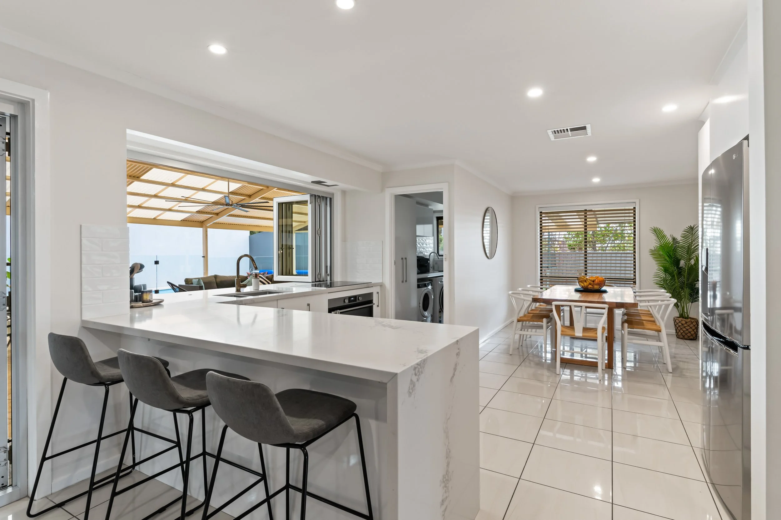 Modern kitchen and dining area with white cabinets, a marble island with three gray chairs, a wooden dining table with white chairs, large window blinds, and a sliding door opening to an outdoor patio with a pergola.
