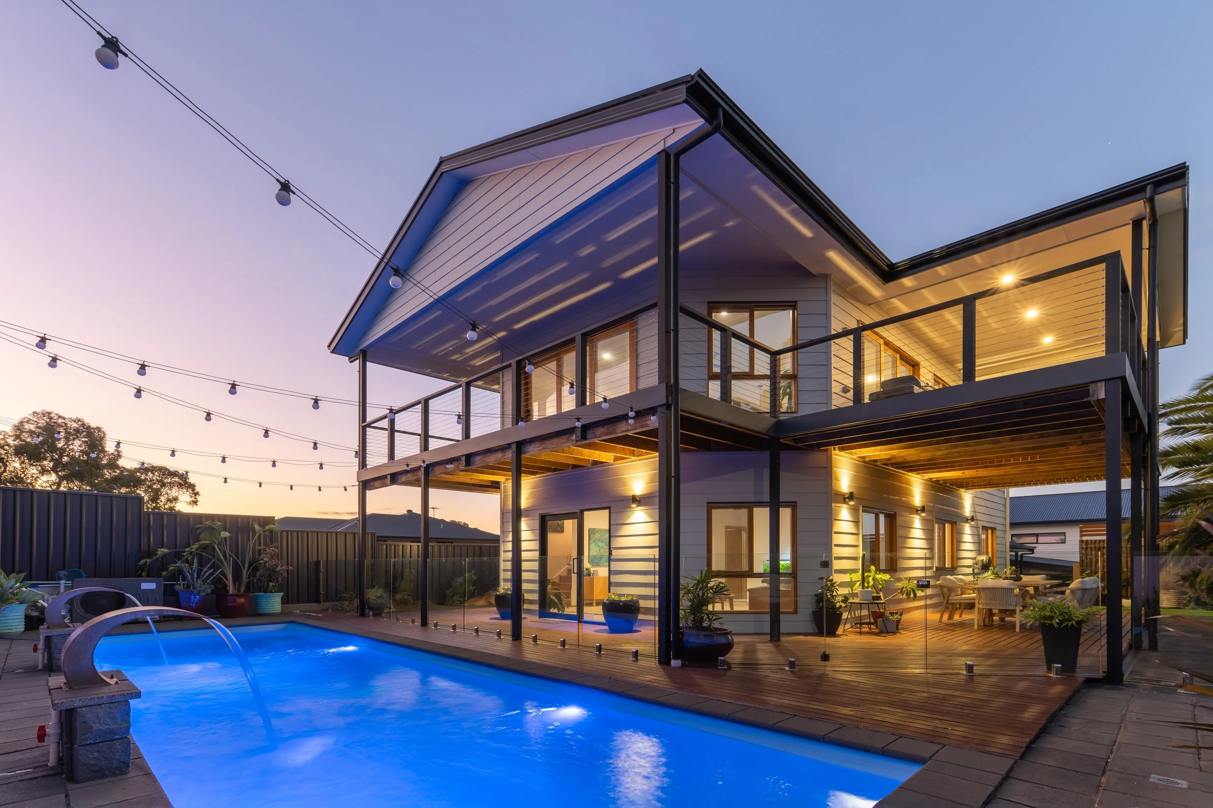 Modern two-story house with a balcony and outdoor patio, illuminated at dusk, with string lights overhead, a swimming pool in the foreground, and trees in the background.