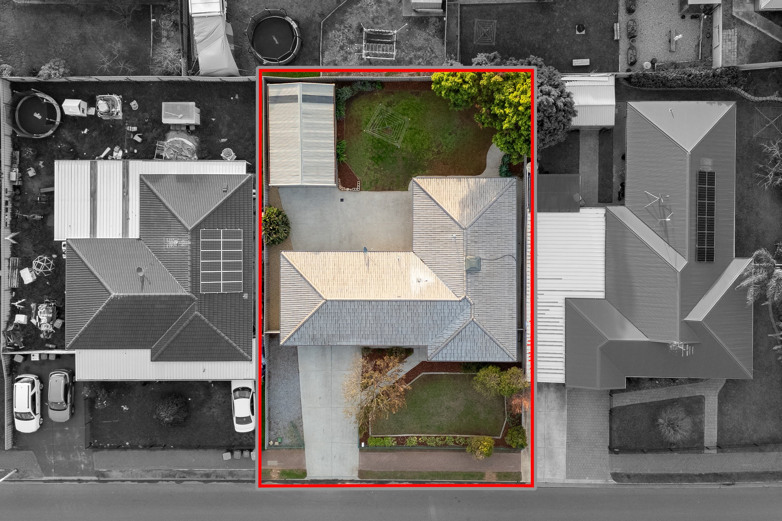 Aerial view of a backyard with a small lawn, a concrete driveway, and neighboring rooftops. The backyard is outlined in red, showing a green lawn, a shed, and a large tree.