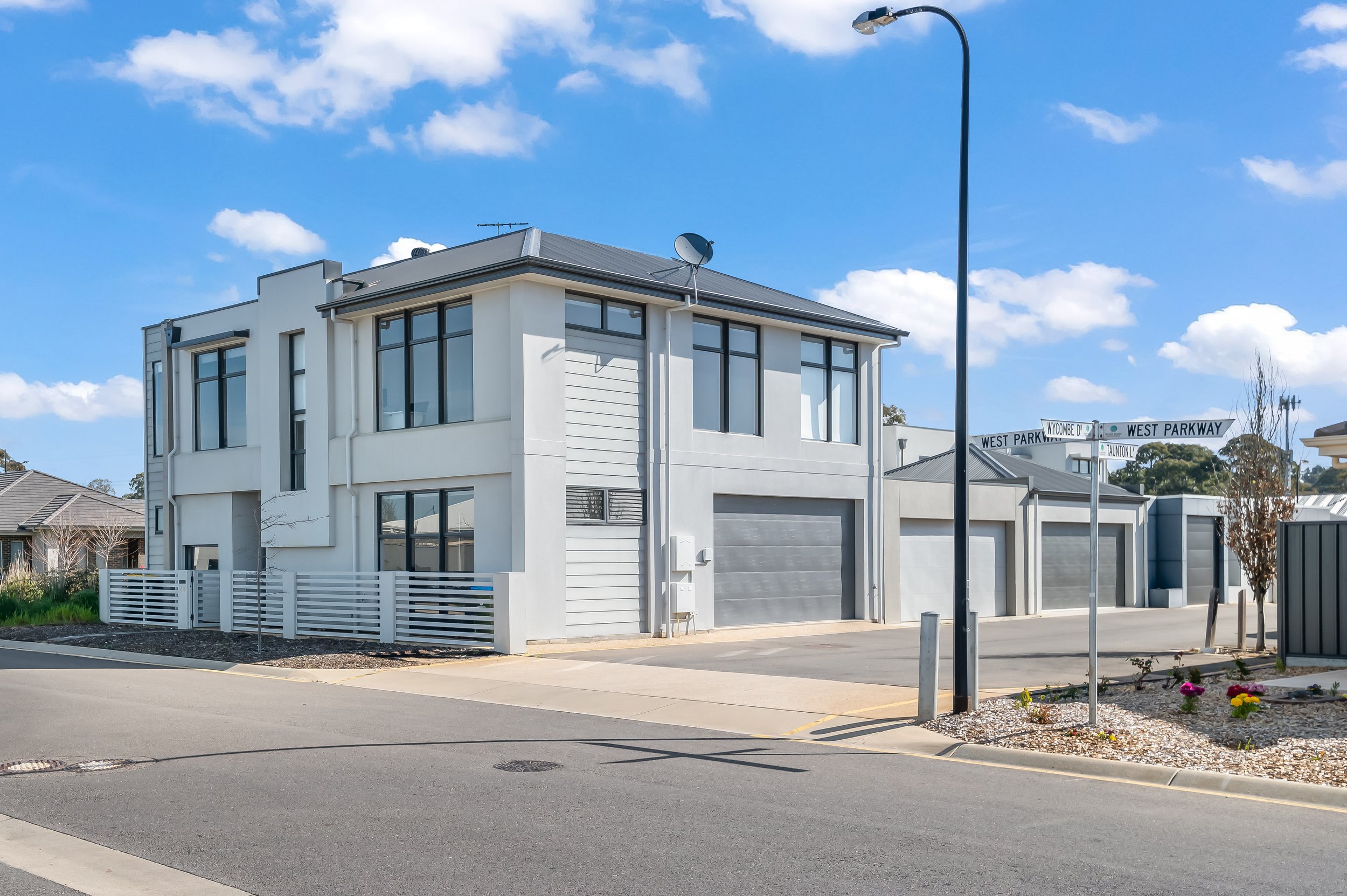 A modern white two-story house with large windows and a gray garage door, situated on a street corner under a blue sky with scattered clouds, near street signs for Wycombe Drive and West Parkway.