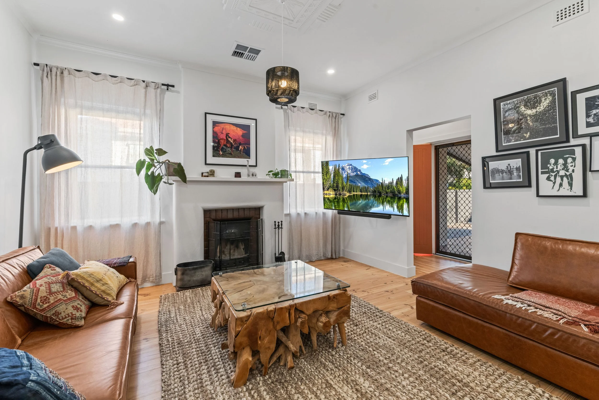 Living room with white walls, a fireplace, and framed artwork, featuring two leather sofas, a glass coffee table, a patterned rug, and a television showing a mountain landscape, with natural light coming through curtains.