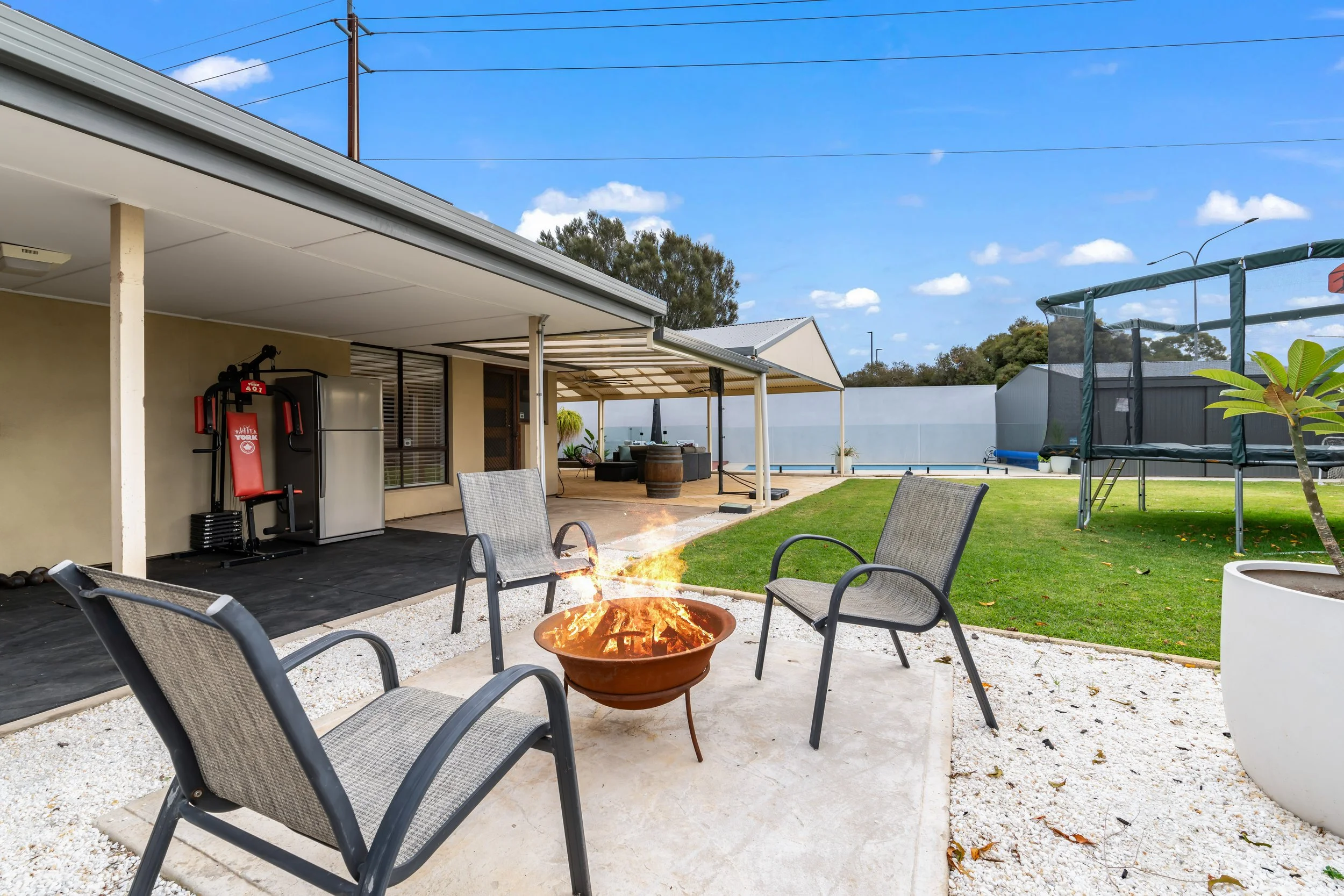 Backyard patio with a fire pit, surrounded by metal chairs, with a house in the background featuring a covered outdoor seating area, a trampoline, and a grassy yard under a clear blue sky.