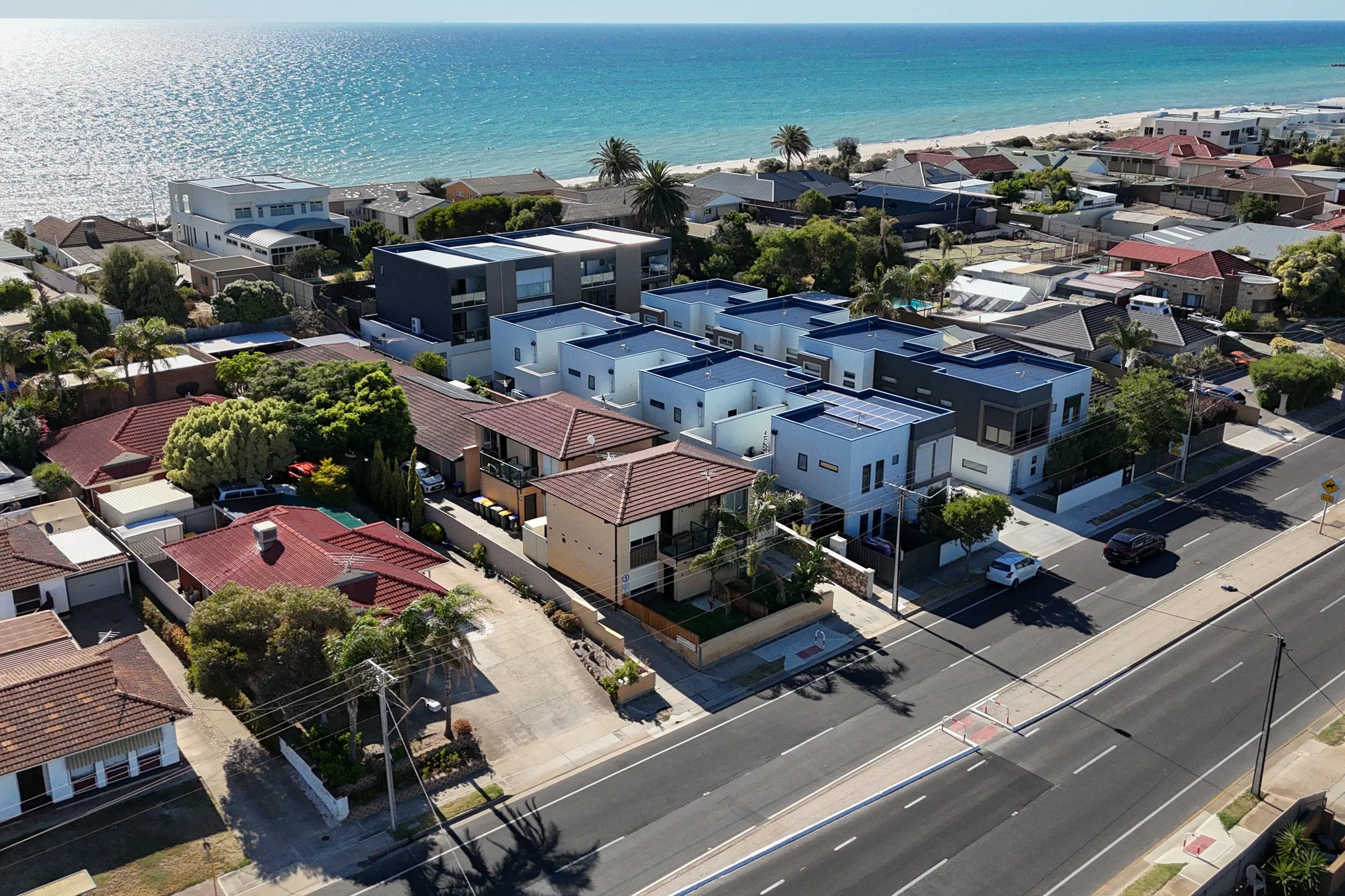 Aerial view of residential neighborhood near a beach with ocean in the background. House rooftops include red, gray, and black tiles, with some modern white and gray buildings and lush green trees.