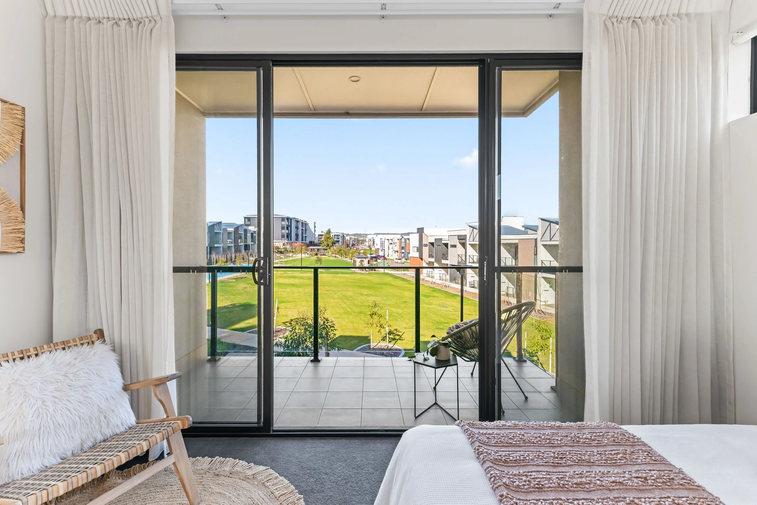 A bedroom with a balcony view of a suburban neighborhood, featuring office chairs, a white bedspread, curtains, a woven chair, and a green lawn outside.