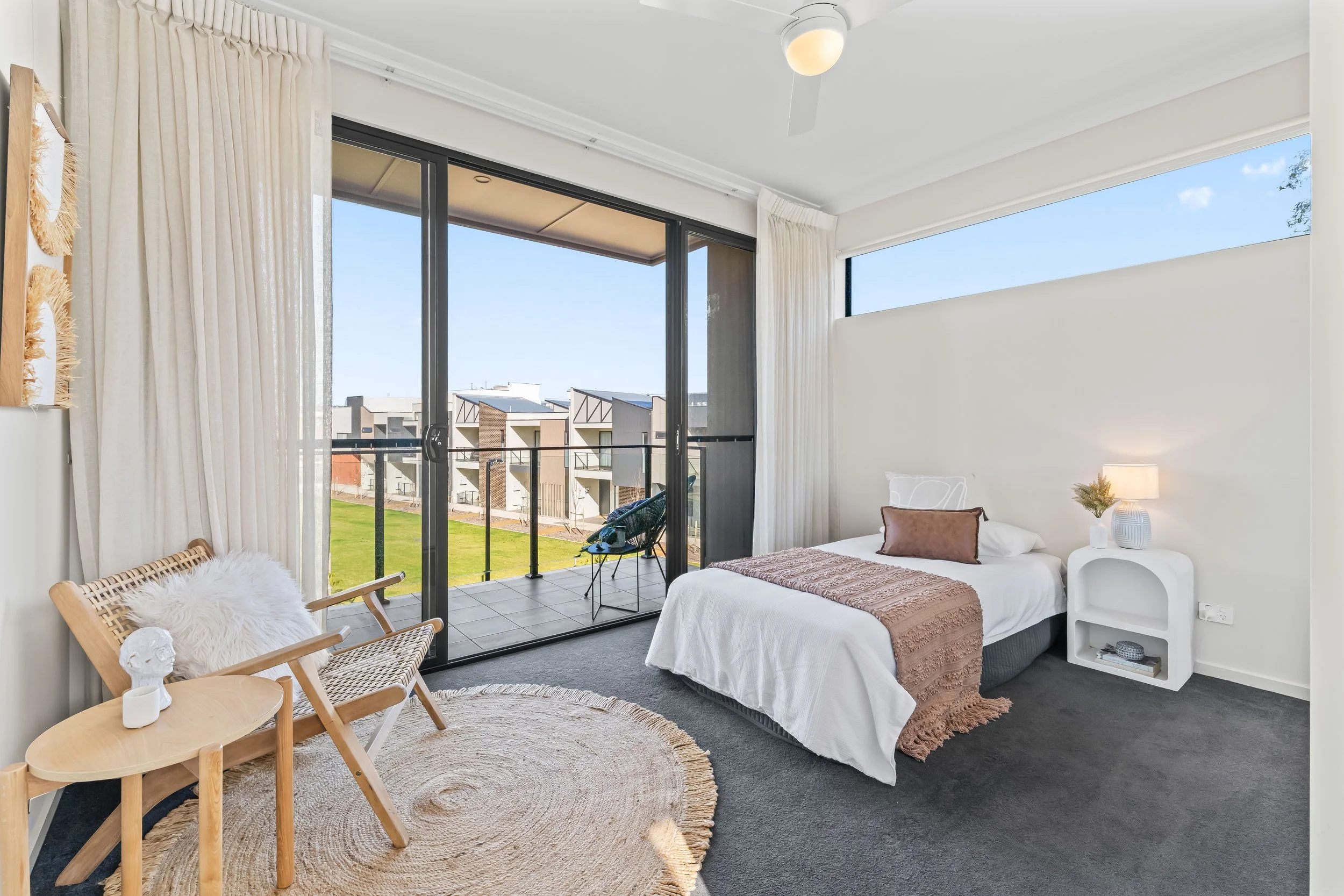 A modern bedroom with a large glass sliding door opening to a balcony, white walls, a bed with white bedding and a brown throw, a small white nightstand with decorative items, a ceiling fan, a patio chair with a fluffy pillow, a circular woven rug, a