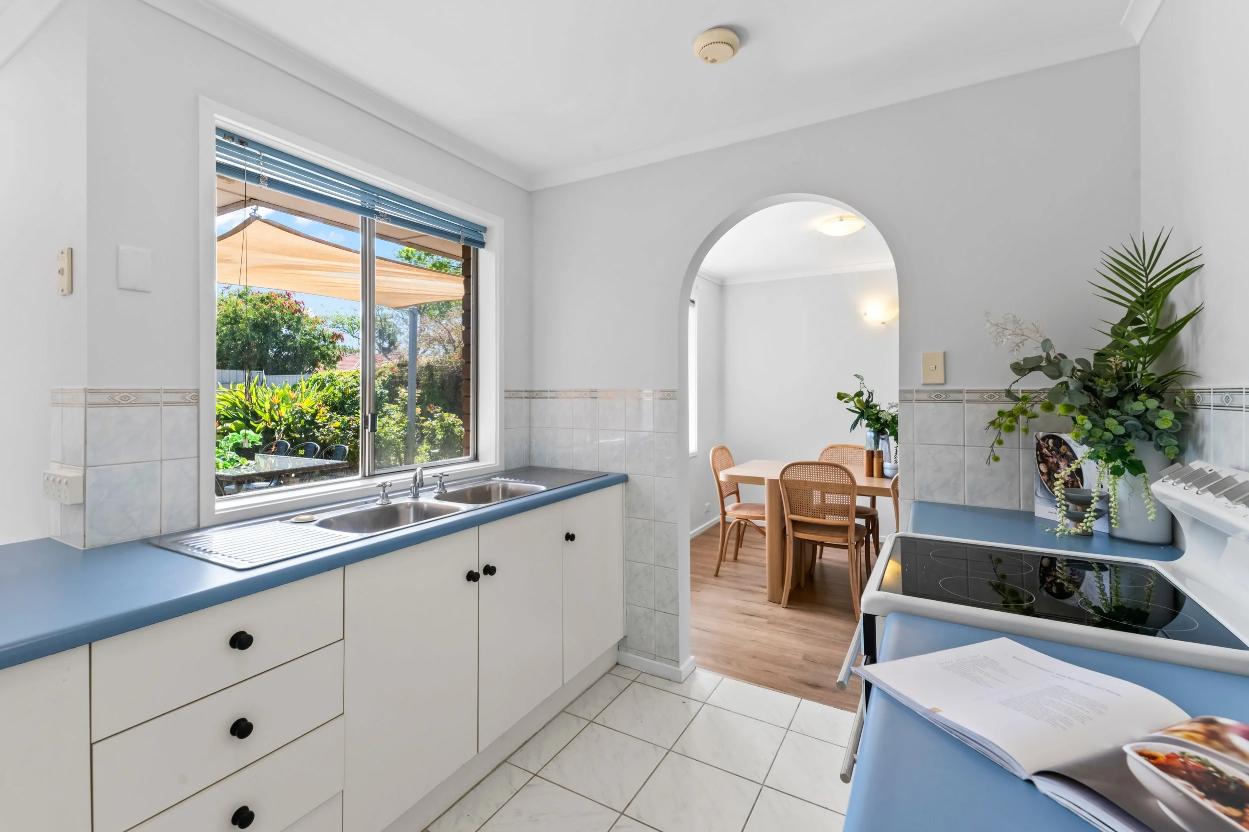 A kitchen with white cabinets and a double sink overlooking a garden through a large window. There is a dining area with a wooden table and chairs, and some potted plants on the side.