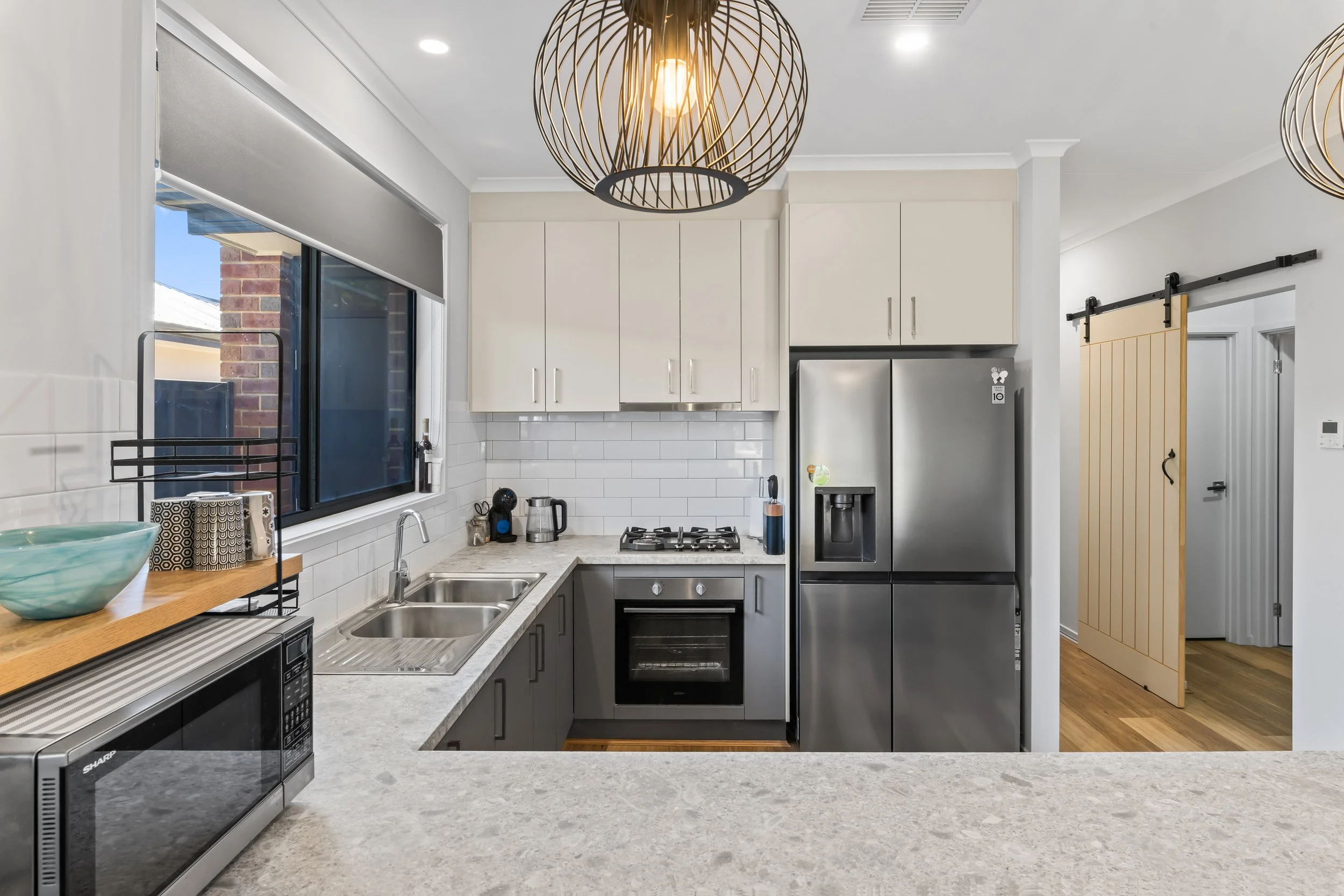 Modern kitchen with white cabinets, stainless steel refrigerator, black stove, microwave, and countertop with various small kitchen appliances and decor, large window with a view of a brick exterior, stylish hanging light fixture, and a sliding barn 