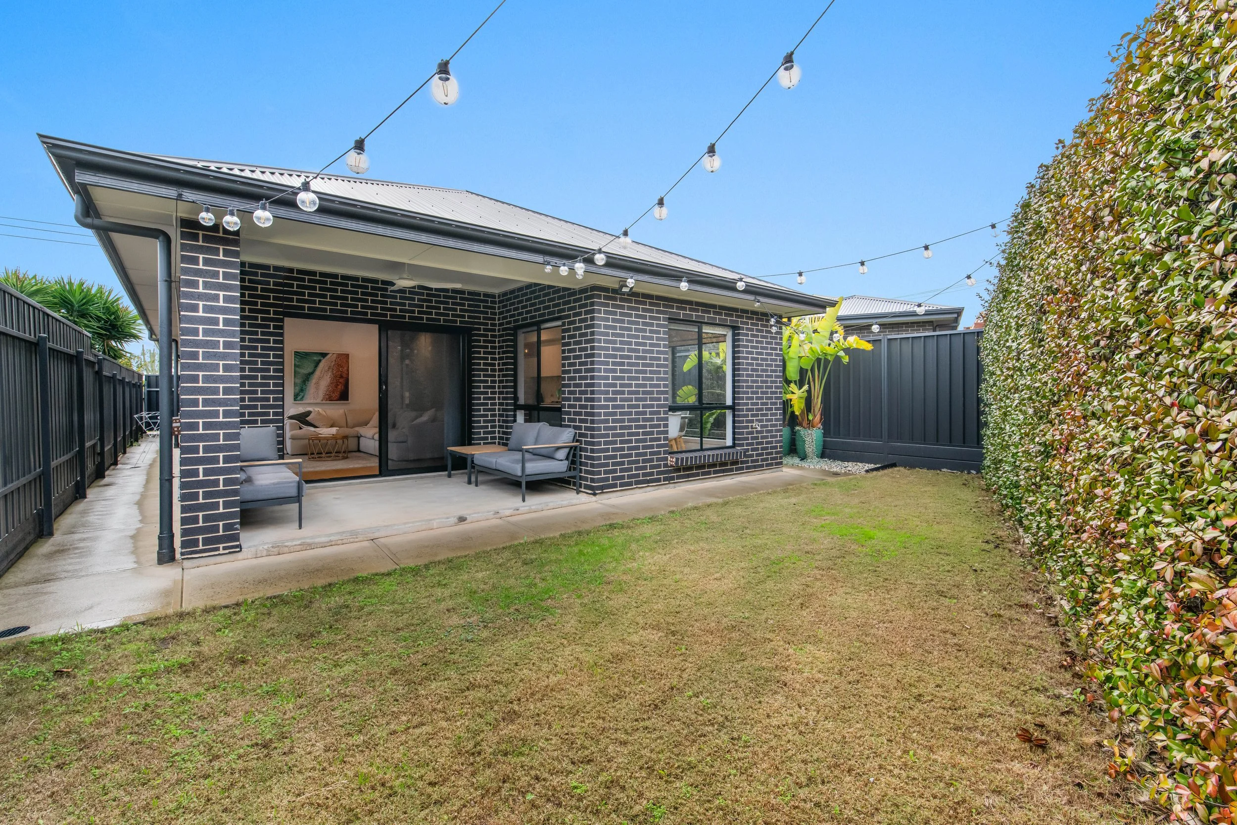 A backyard patio with two gray outdoor chairs, a small table, and a sliding glass door leading to an indoor living room. The patio is covered with string lights and surrounded by a black metal fence and hedges. There is a small patch of grass in the 
