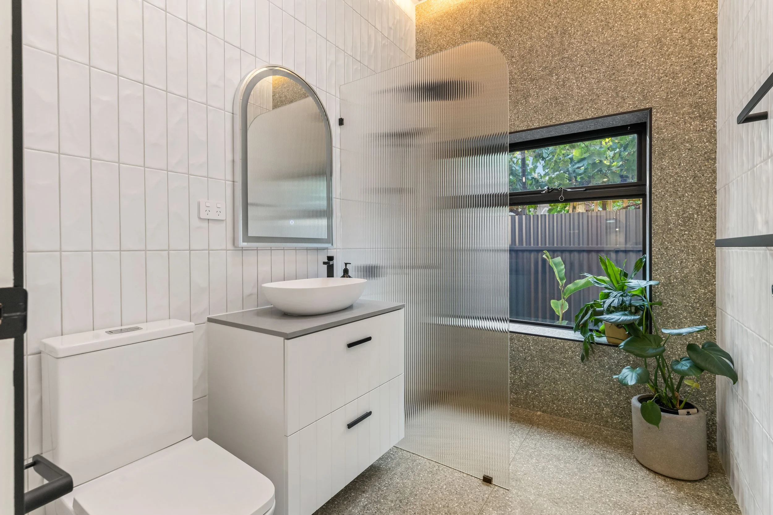 A modern bathroom with white tiled walls, a white toilet, a white vanity with a vessel sink, a curved mirror, a window with a view of greenery outside, and green plants in pots. There is a textured concrete wall on one side and a frosted glass privac
