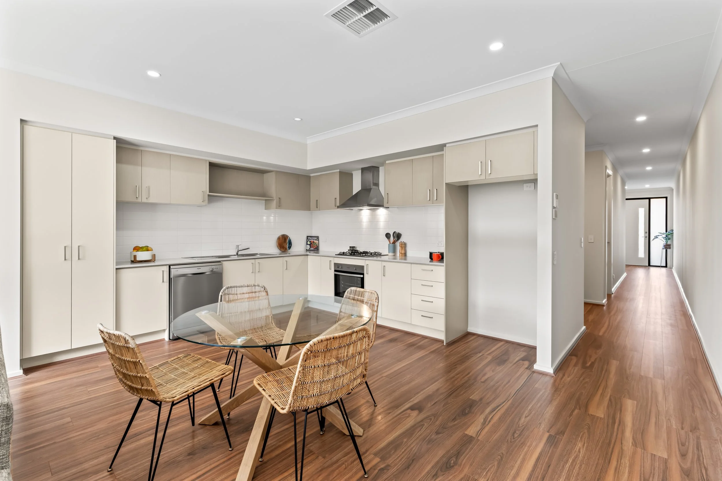 Modern kitchen with white cabinets, stainless steel appliances, and a glass dining table surrounded by wicker chairs, hardwood floors, and a hallway in the background.