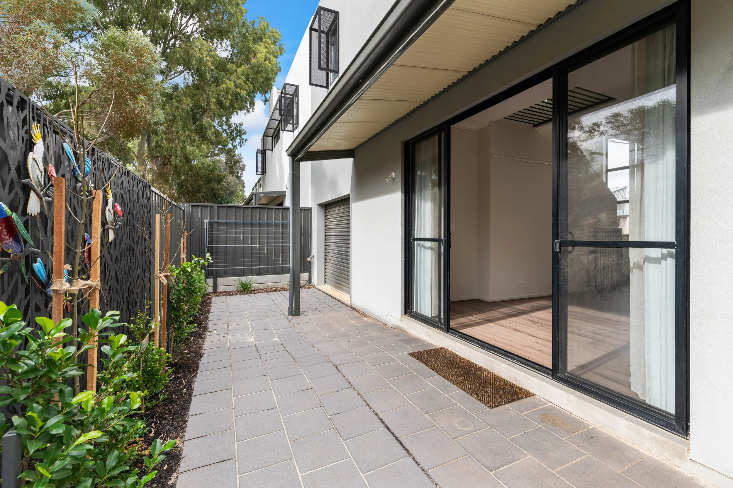 Modern house exterior with sliding glass doors leading to an outdoor patio, gray tiled flooring, and a decorative fence with colorful bird ornaments, surrounded by green plants and trees under a partly cloudy sky.
