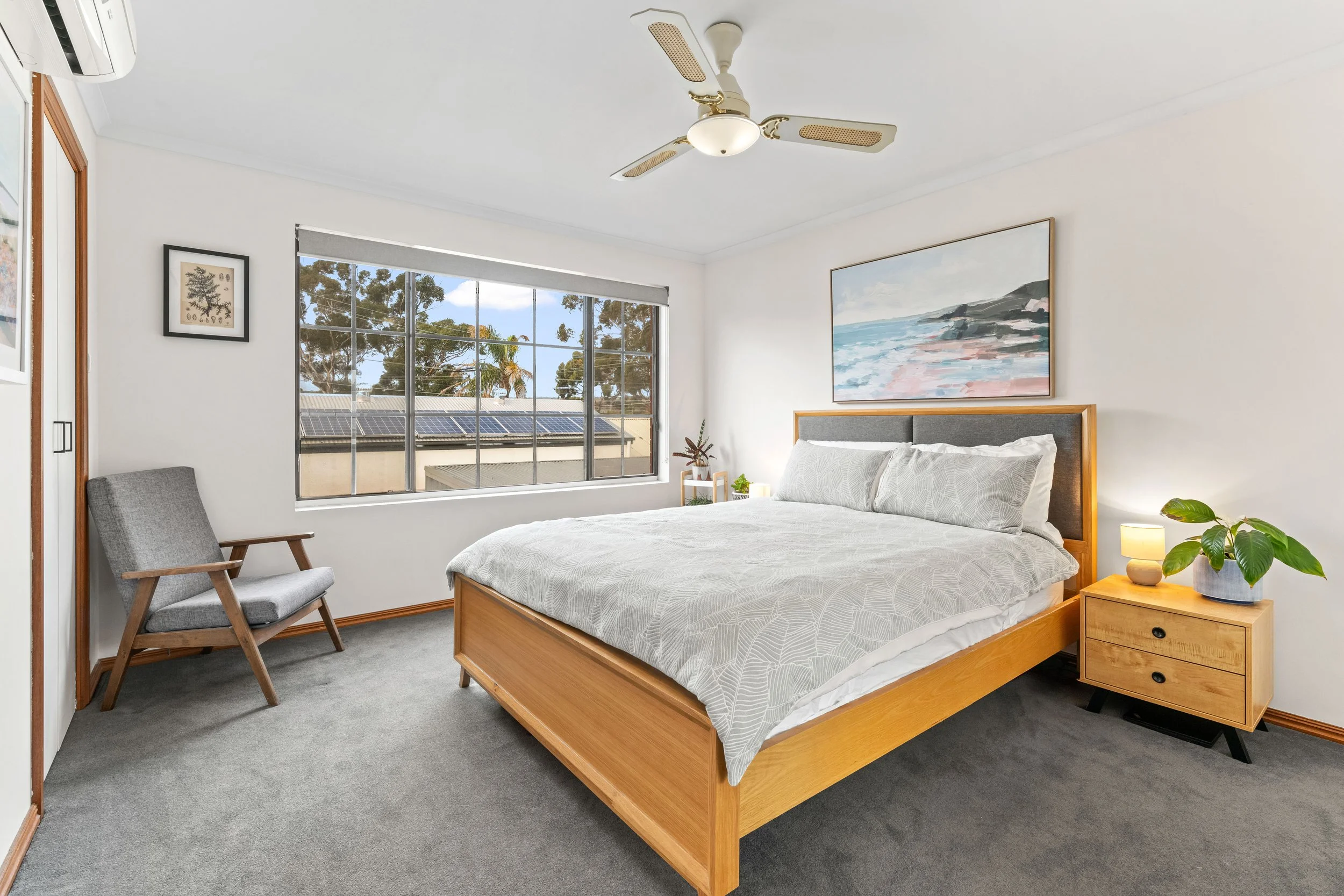 A bedroom with white walls and a large window showing trees outside. There is a wooden bed with light-colored bedding, a nightstand with a lamp and potted plant, a gray upholstered chair, and a framed artwork of a seascape above the bed. A ceiling fa