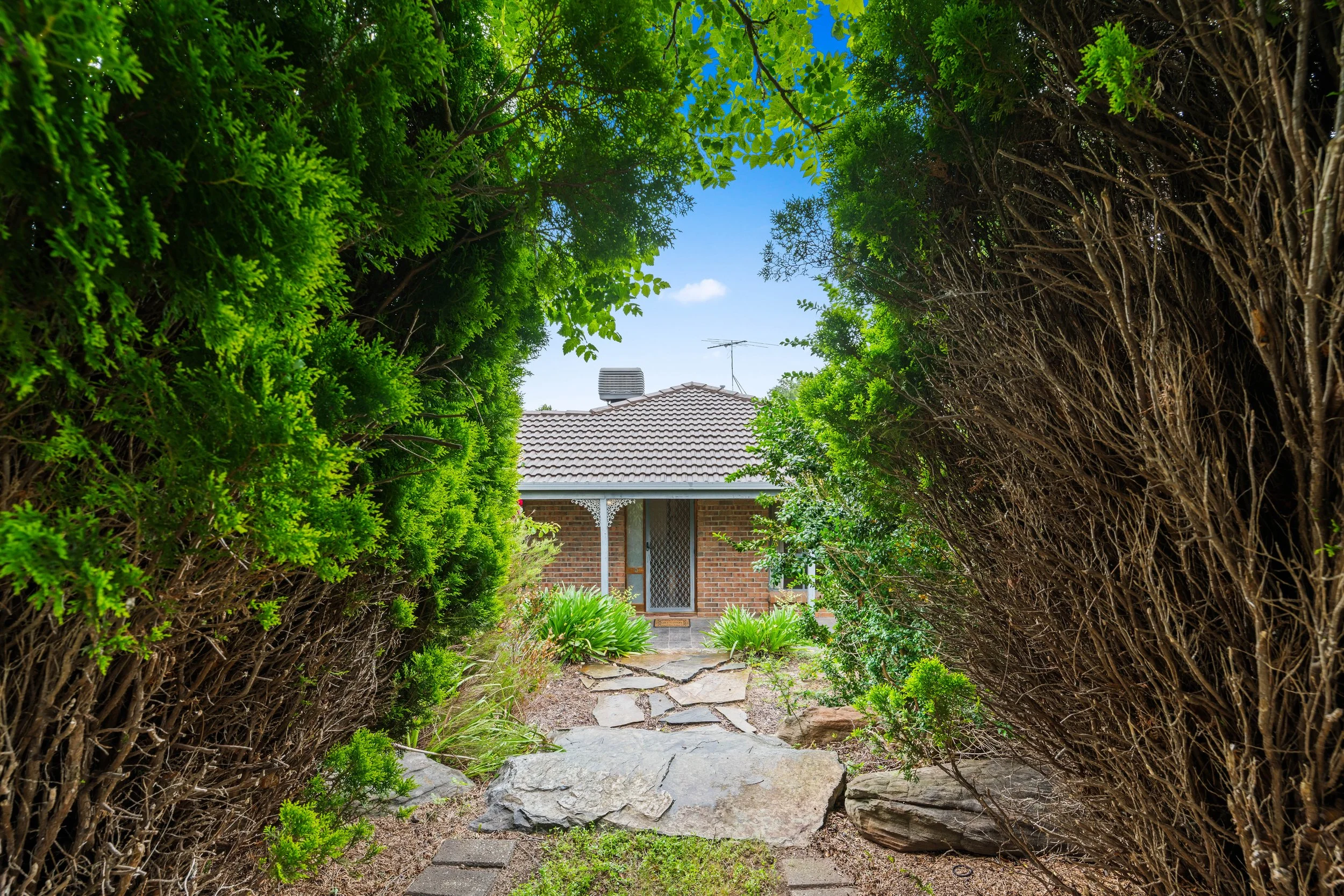 View through a hedge archway leading to a brick house with a tiled roof, greenery, and a stone pathway.