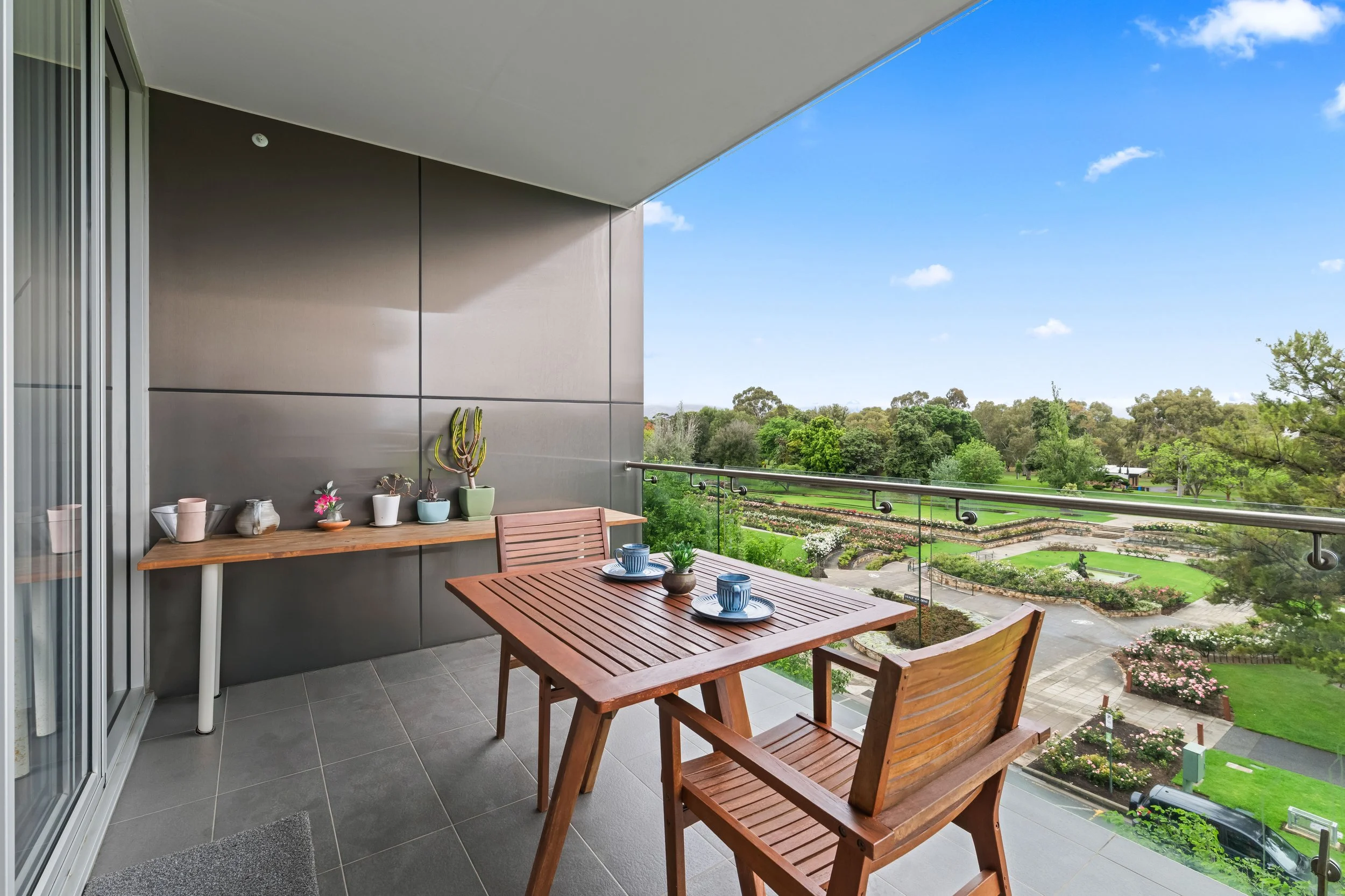 Balcony with a wooden table set with cups and small plants, overlooking a green park with trees and garden beds under a blue sky with a few clouds.