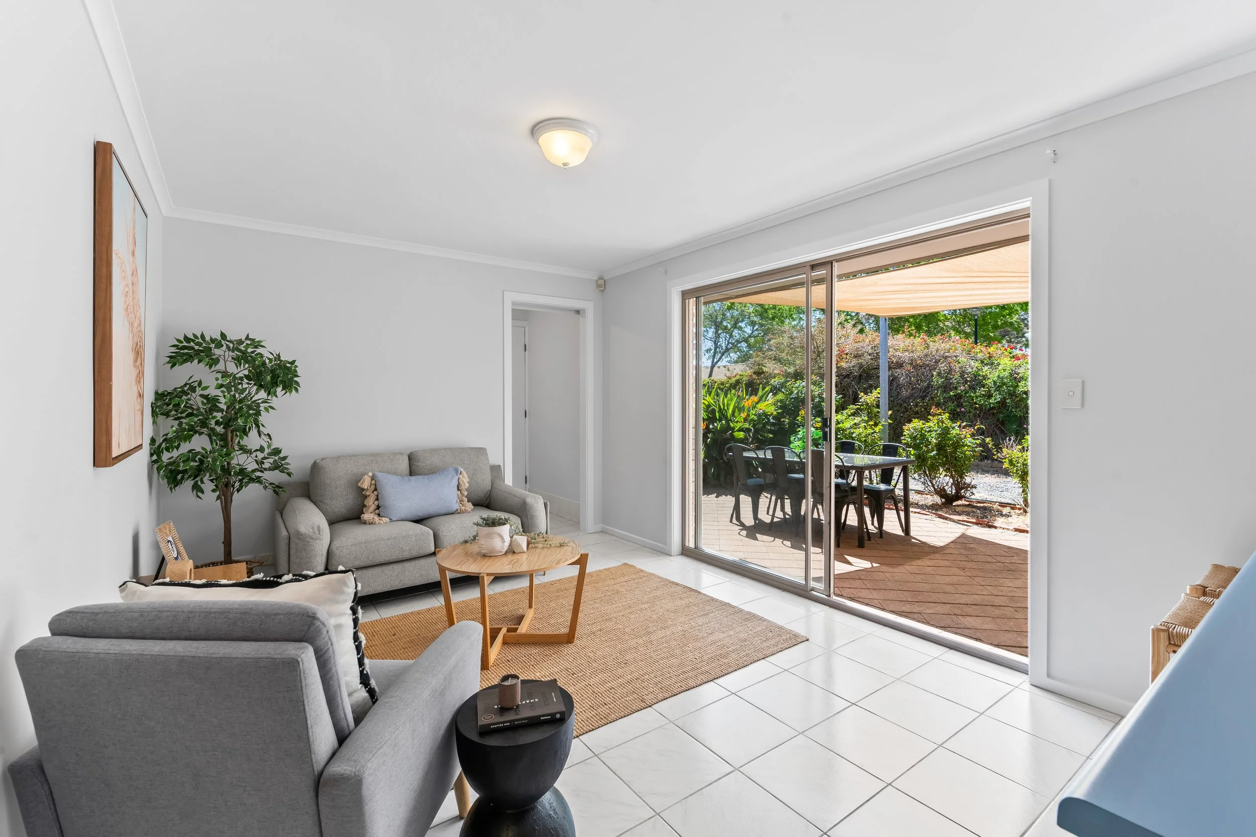 Living room with gray couch, potted plant, wooden coffee table, sliding glass door leading to outdoor patio with dining table and chairs, garden view.