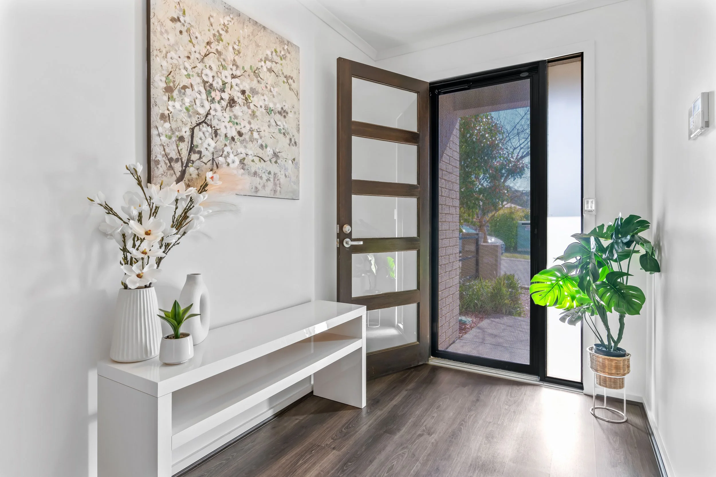 A minimalist entryway with a white console table holding white vases with flowers, and potted plants including a monstera, next to a screen door and a wall with a large floral painting.