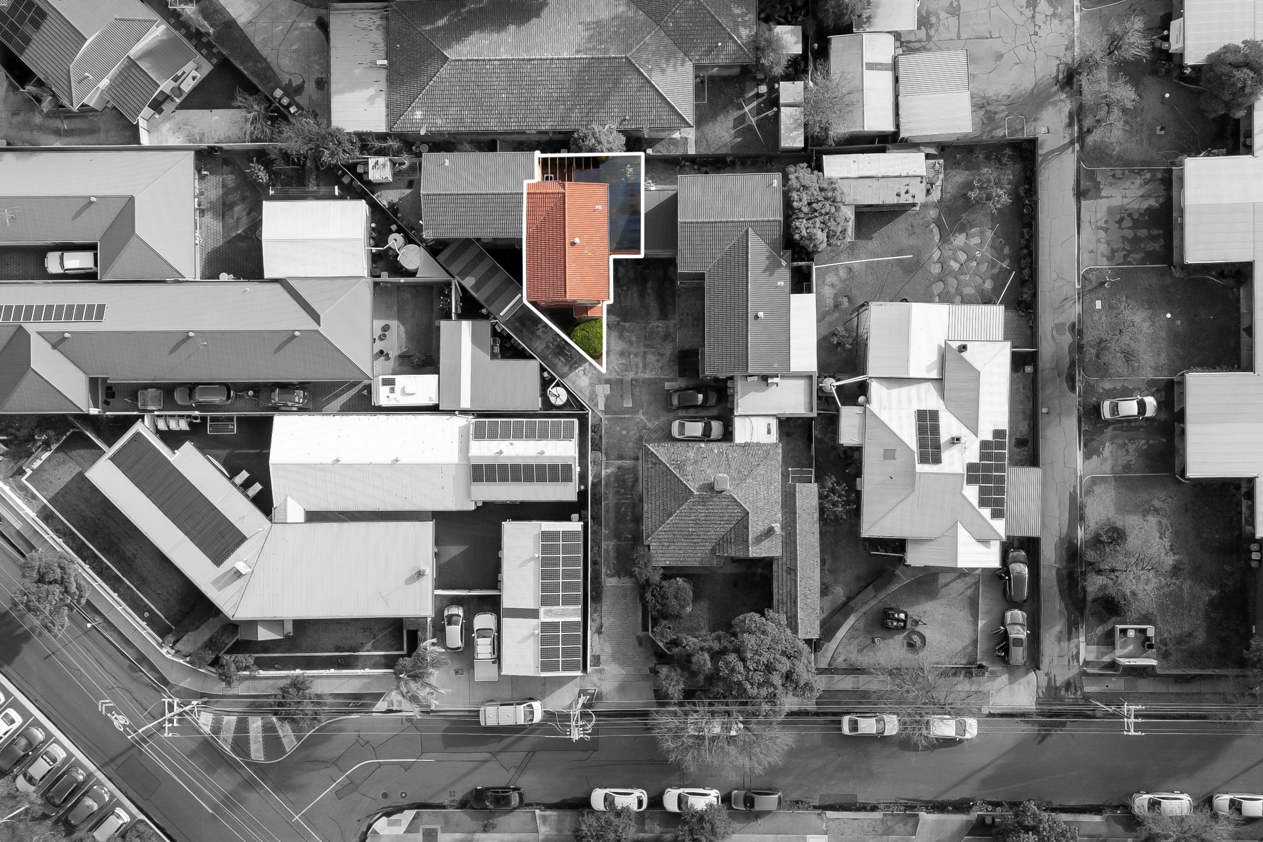 An aerial view of a residential neighborhood showing multiple houses with different roof styles, parked cars on the streets and in driveways, trees, and utility poles.