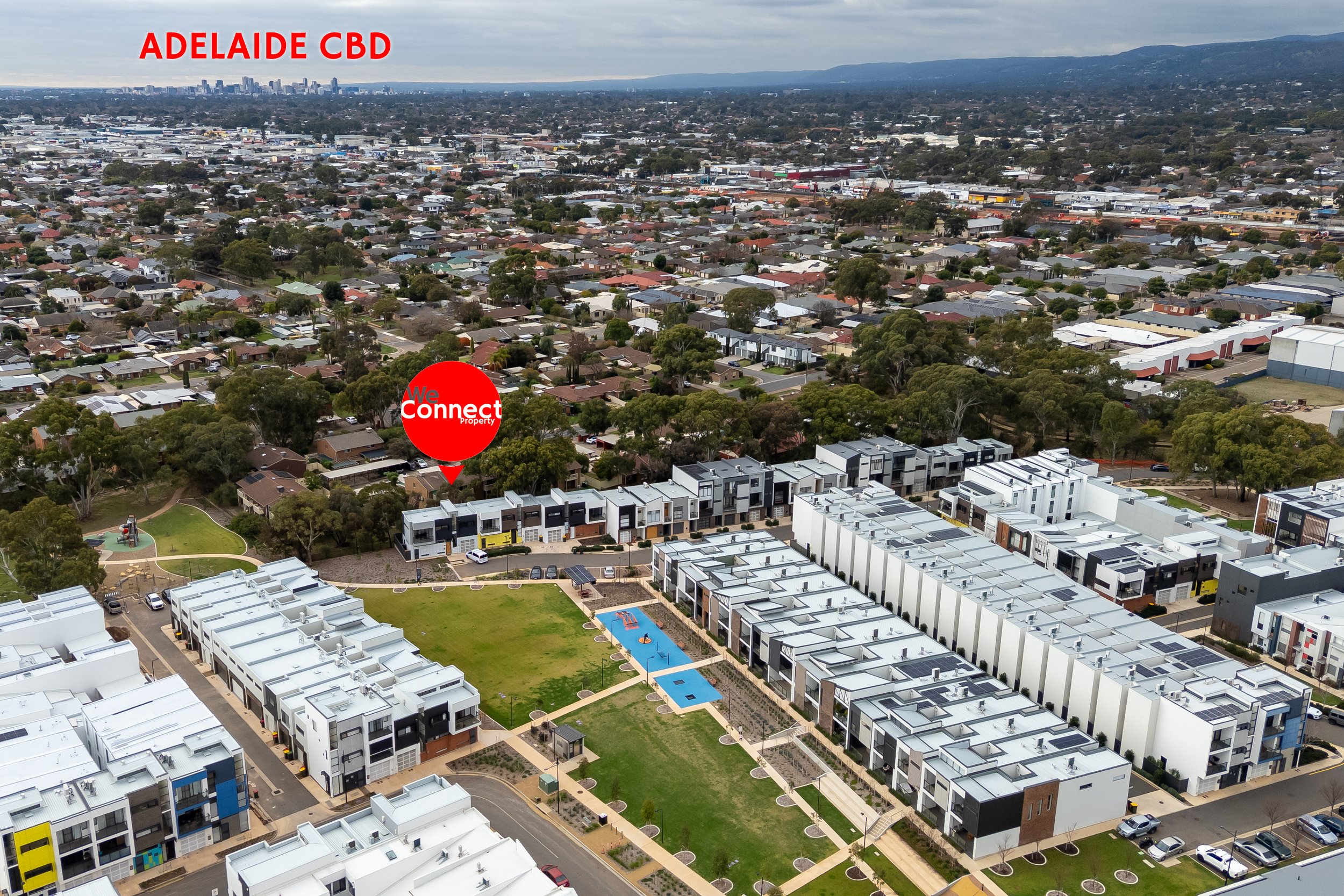 Aerial view of a city with residential neighborhoods, commercial areas, and a park with a swimming pool. The image highlights Adelaide CBD in the background, and a property with a red marker labeled 'We Connect Property' in a suburban area.