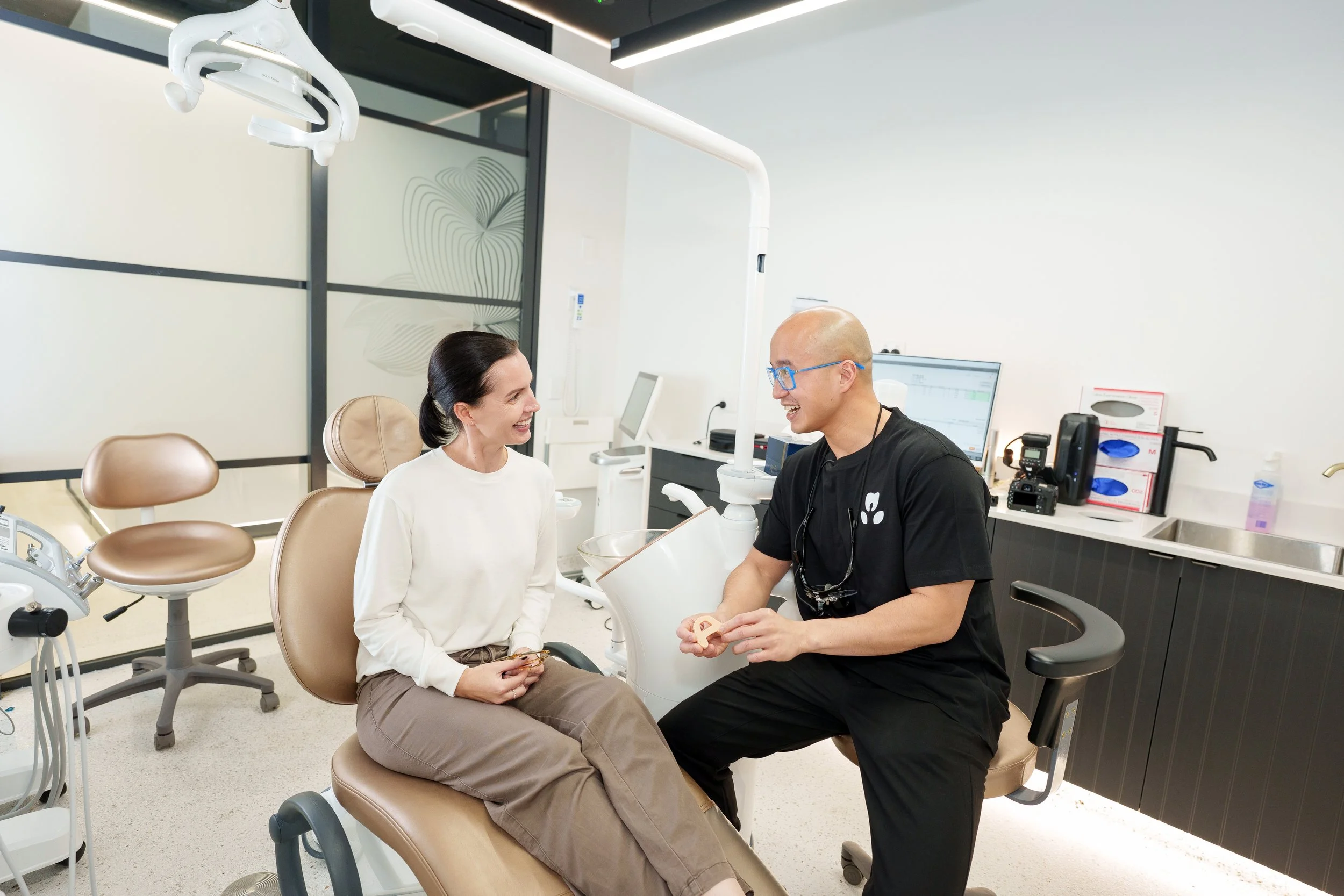 A woman sitting in a dental chair smiling and talking with a male dentist who is wearing glasses and a black shirt, in a modern dental clinic.