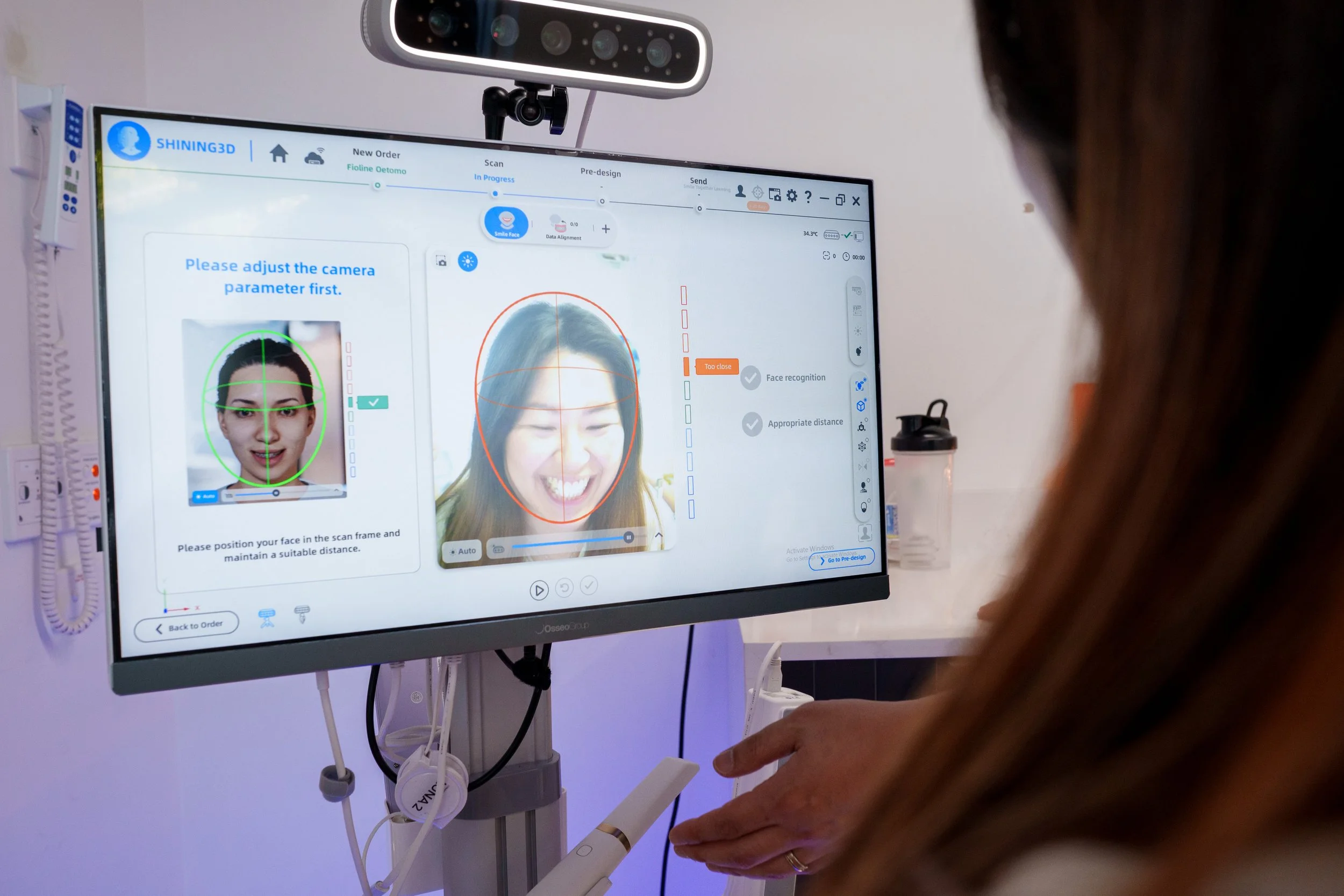 A person is using a computer monitor to scan a face for digital smile design services. The screen displays instructions to position the face within a frame, with a green circle around the face on the left and a red circle on the right.