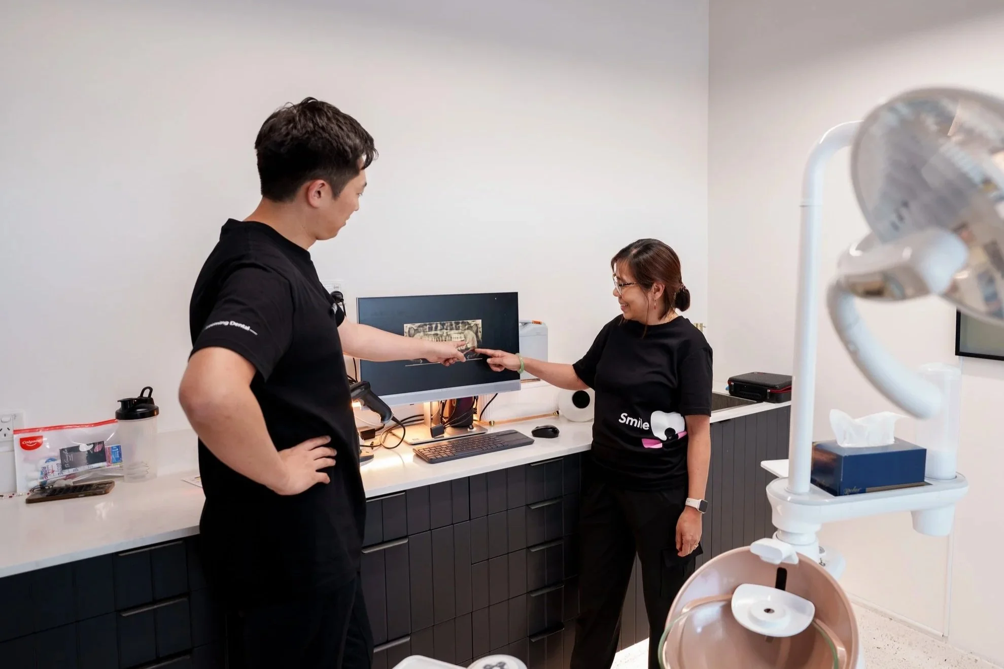 Two people in a medical or dental office exchanging a small item, possibly a card or a check, with one person extending their hand and the other smiling while holding the item. The room has a white counter with various items and medical equipment including a robotic arm, computer monitor, and dental tools.