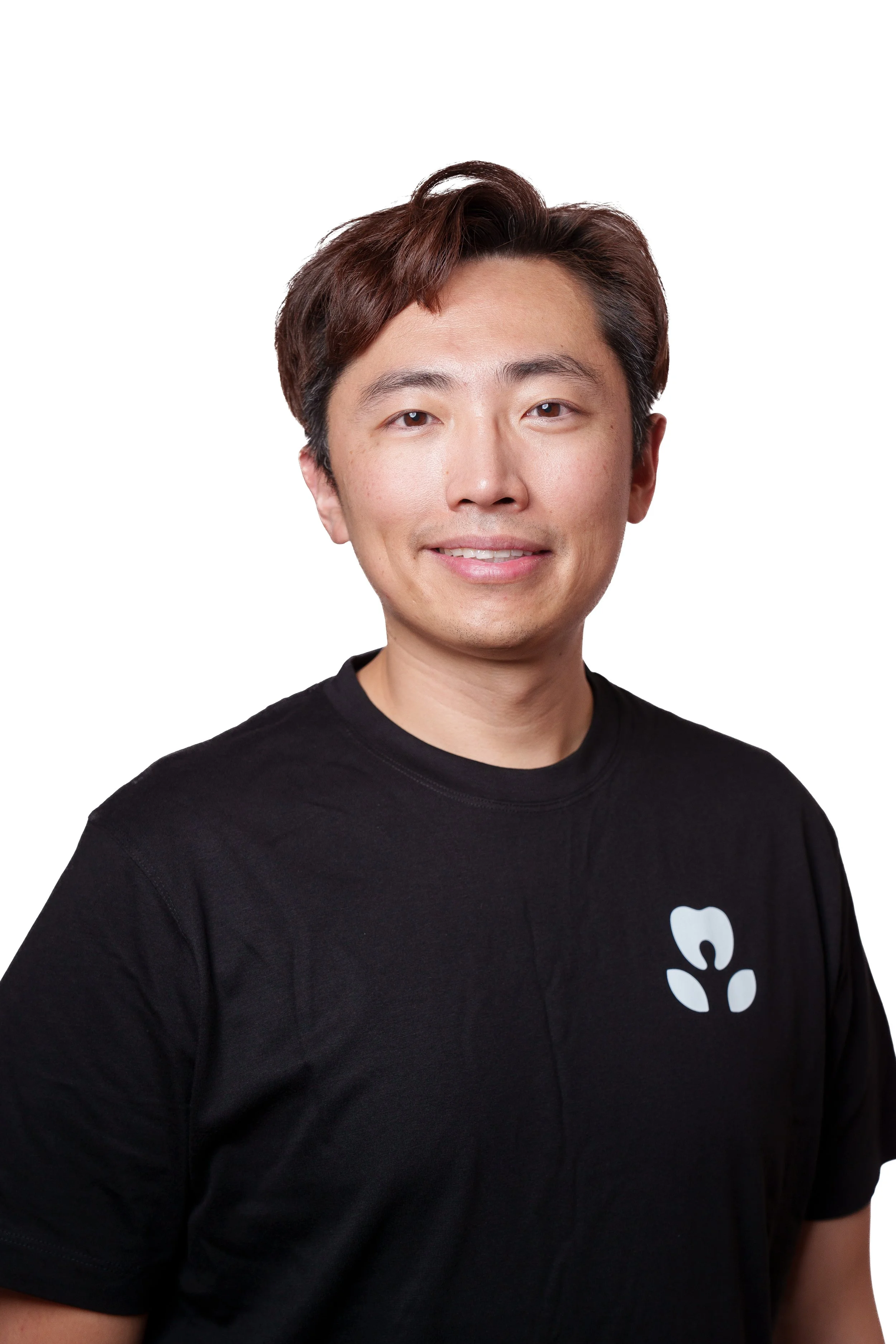 A young man with short brown hair and light skin smiling, wearing a black T-shirt with a white logo on the chest, against a plain white background.