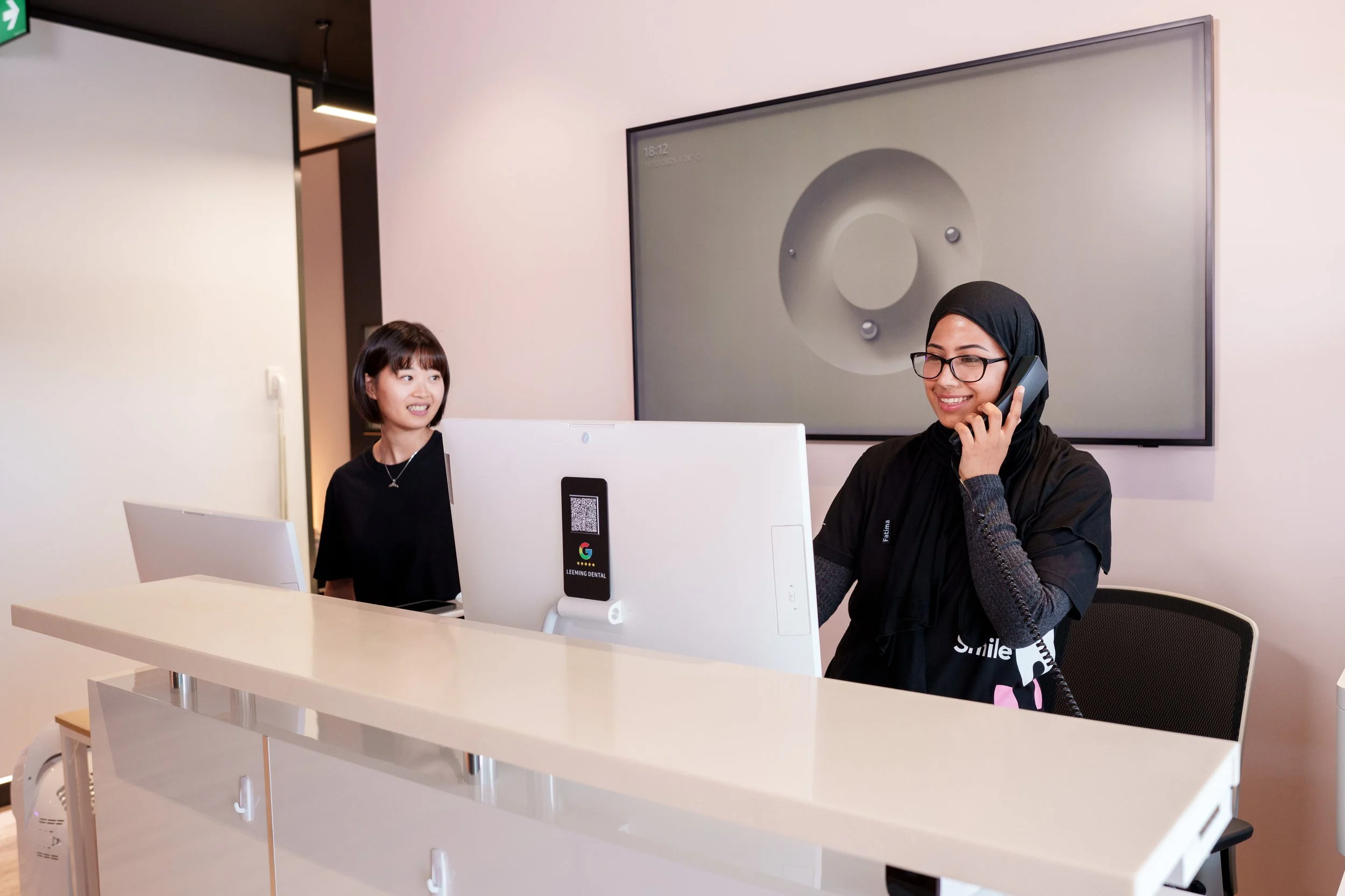 Receptionist smiling while on the phone at a dental clinic front desk, with a patient woman smiling in the background.