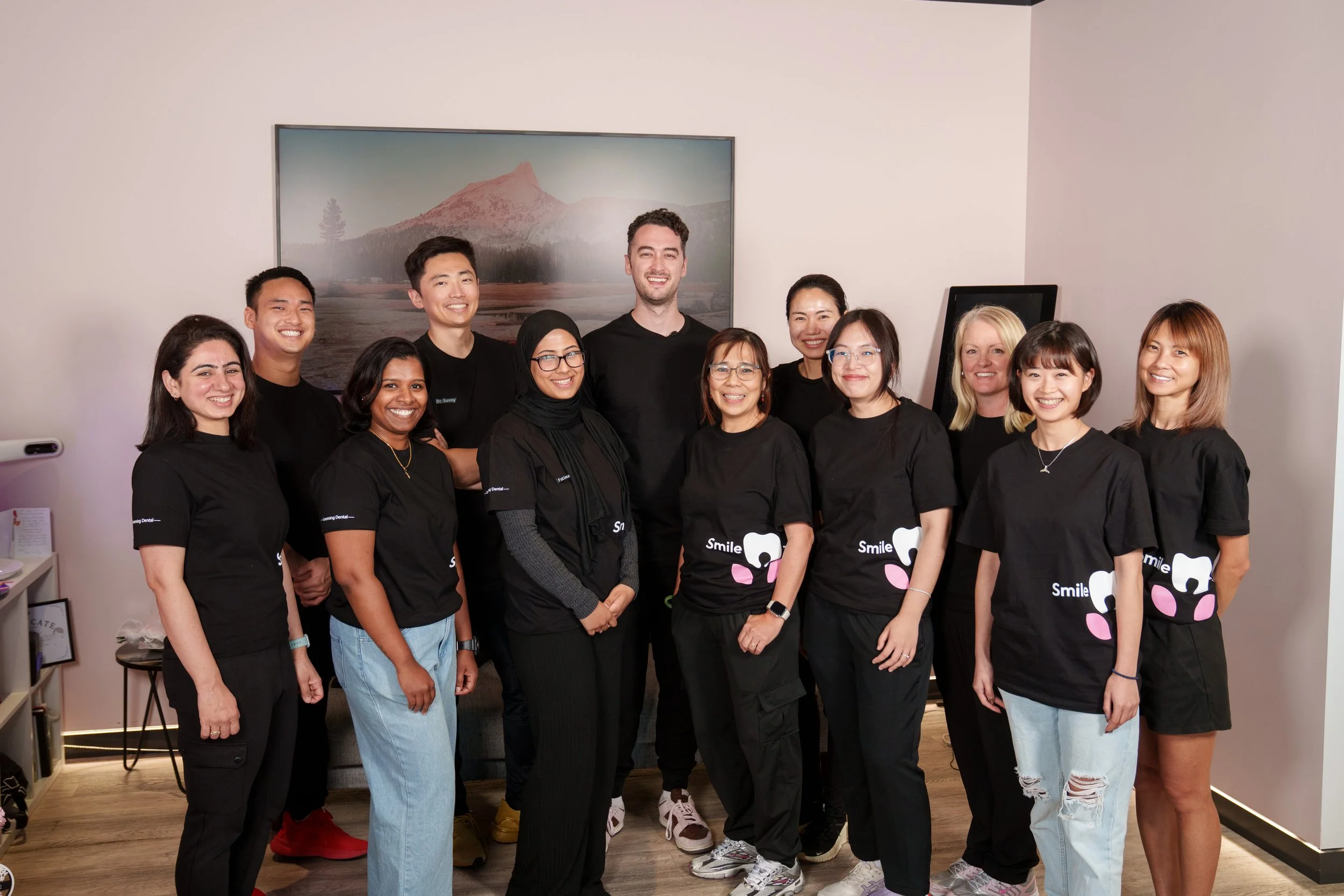 A diverse group of eleven people standing together indoors, smiling at the camera, with a mountain landscape photograph on the wall behind them.