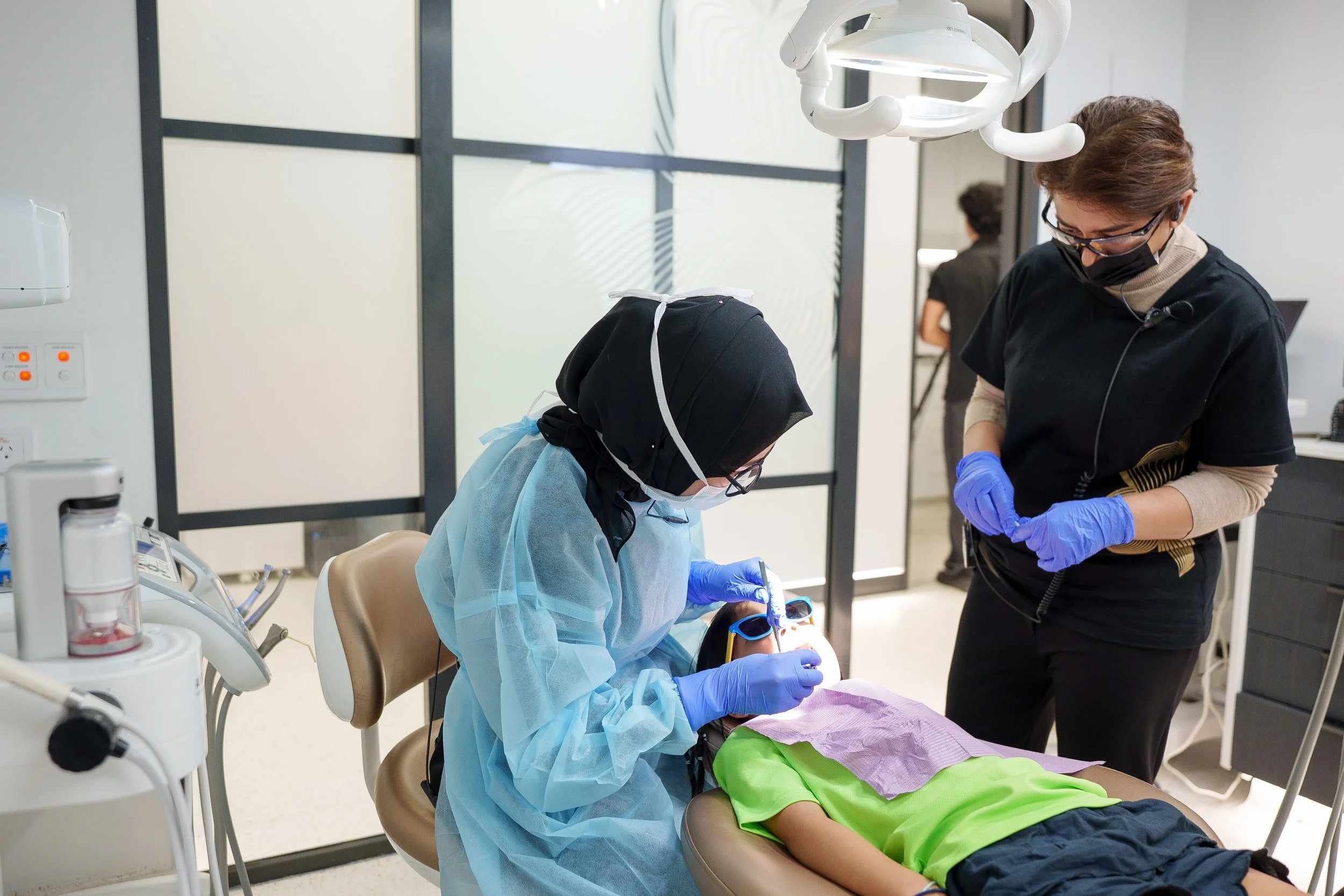 A dentist or dental technician performing a procedure on a patient lying in a dental chair, with dental tools and equipment visible, in a clinical setting.