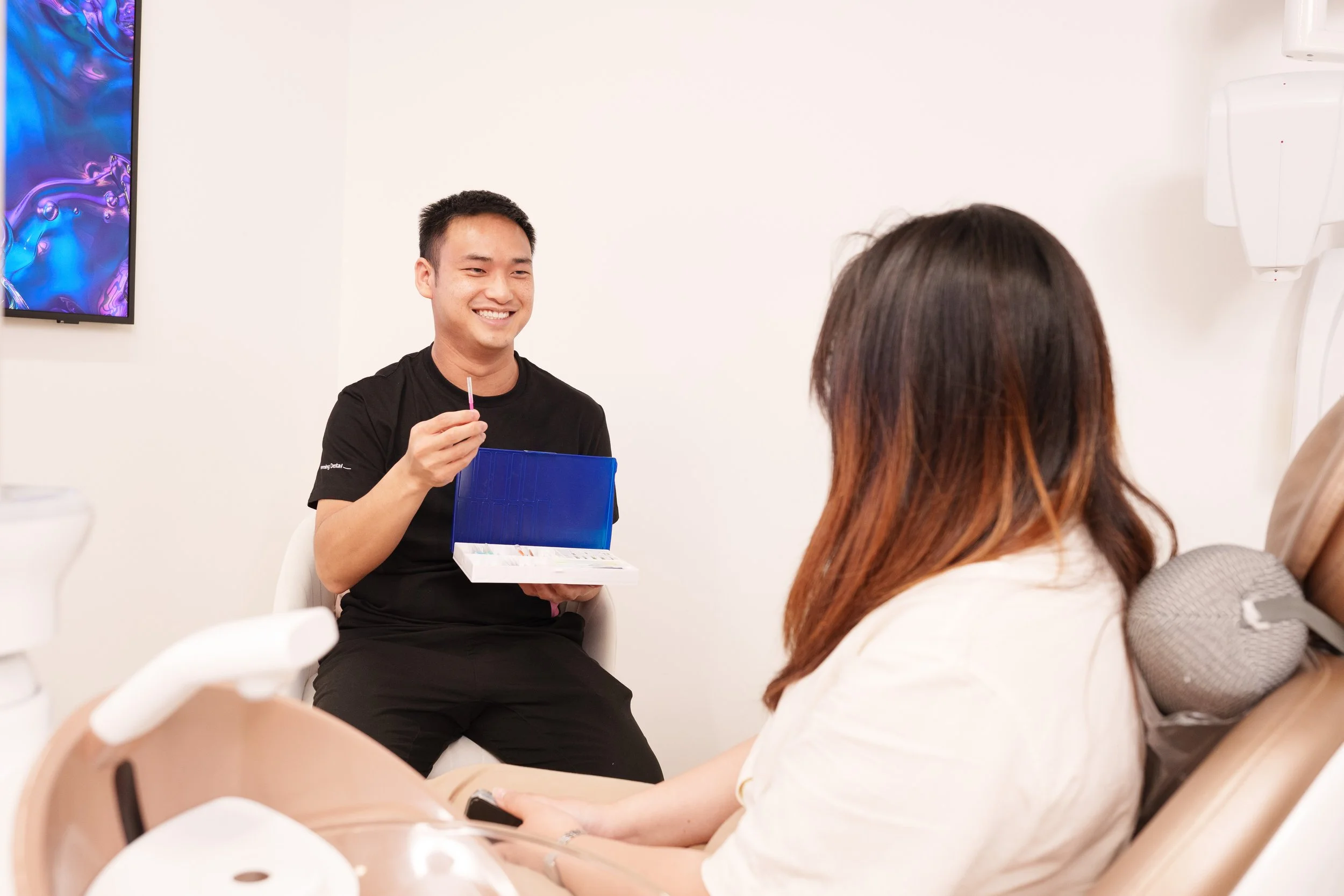 A dental professional sitting and smiling while holding a case of dental tools in front of a patient in a dental clinic.