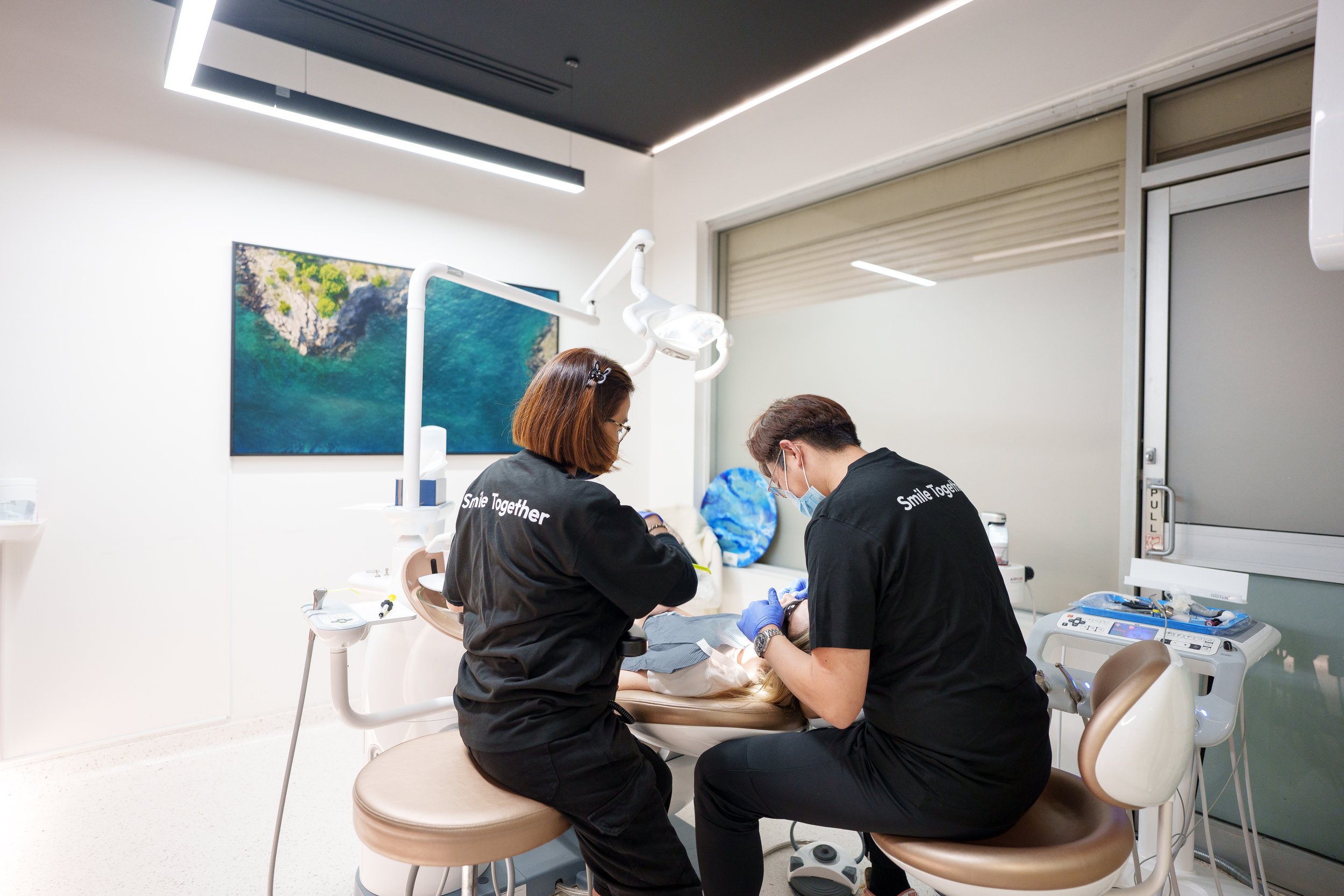 Two dental professionals working on a patient in a dental clinic, with dental equipment, a painting of a landscape, and a window in the background.