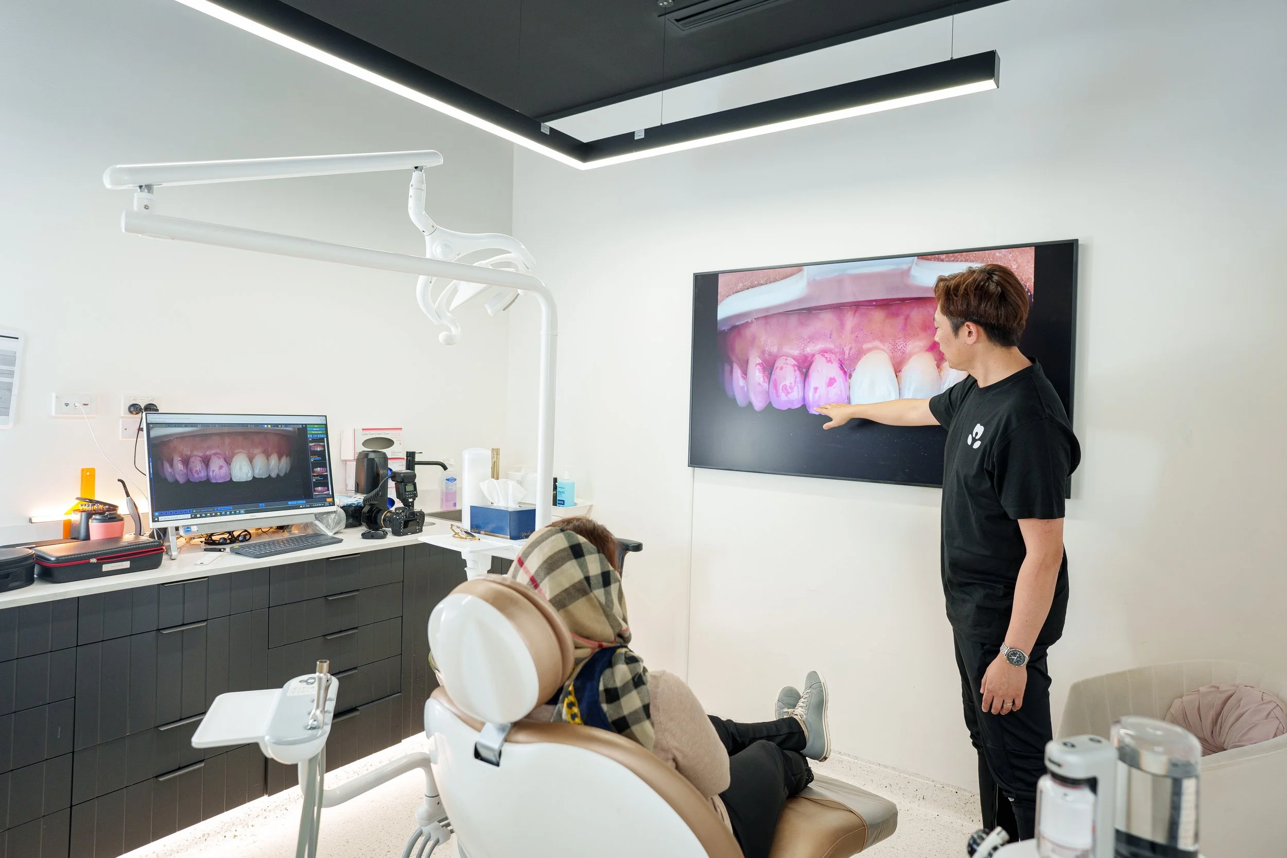 A dental office with a nurse or doctor presenting a digital 3D model of teeth on a large wall-mounted screen. A patient is seated in a dental chair, looking at the screen, with the back of their head visible. The office is modern, with dental equipment and a computer on the counter.