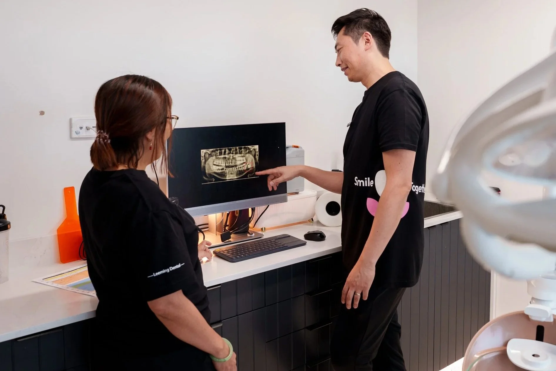 A male dental professional explains a dental X-ray to a female patient in a clinic, both standing near a computer monitor displaying the X-ray.