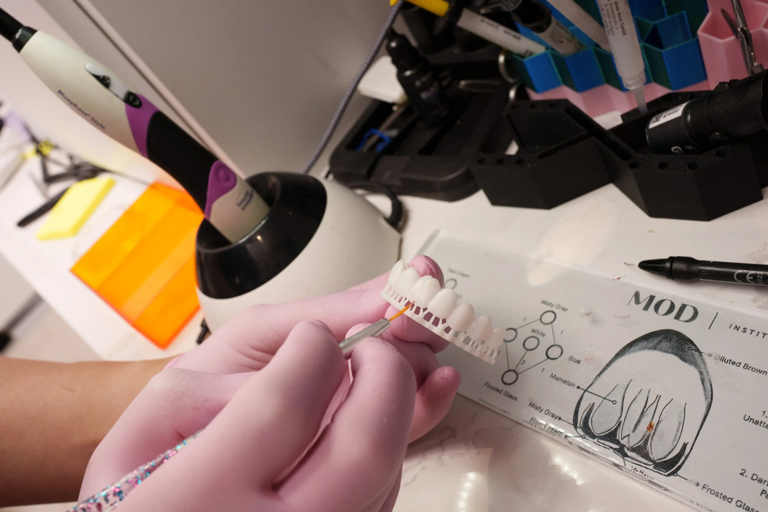 Close-up of a person wearing pink gloves holding a dental shade guide and using a small tool to match teeth color, with dental supplies and a drill in the background.