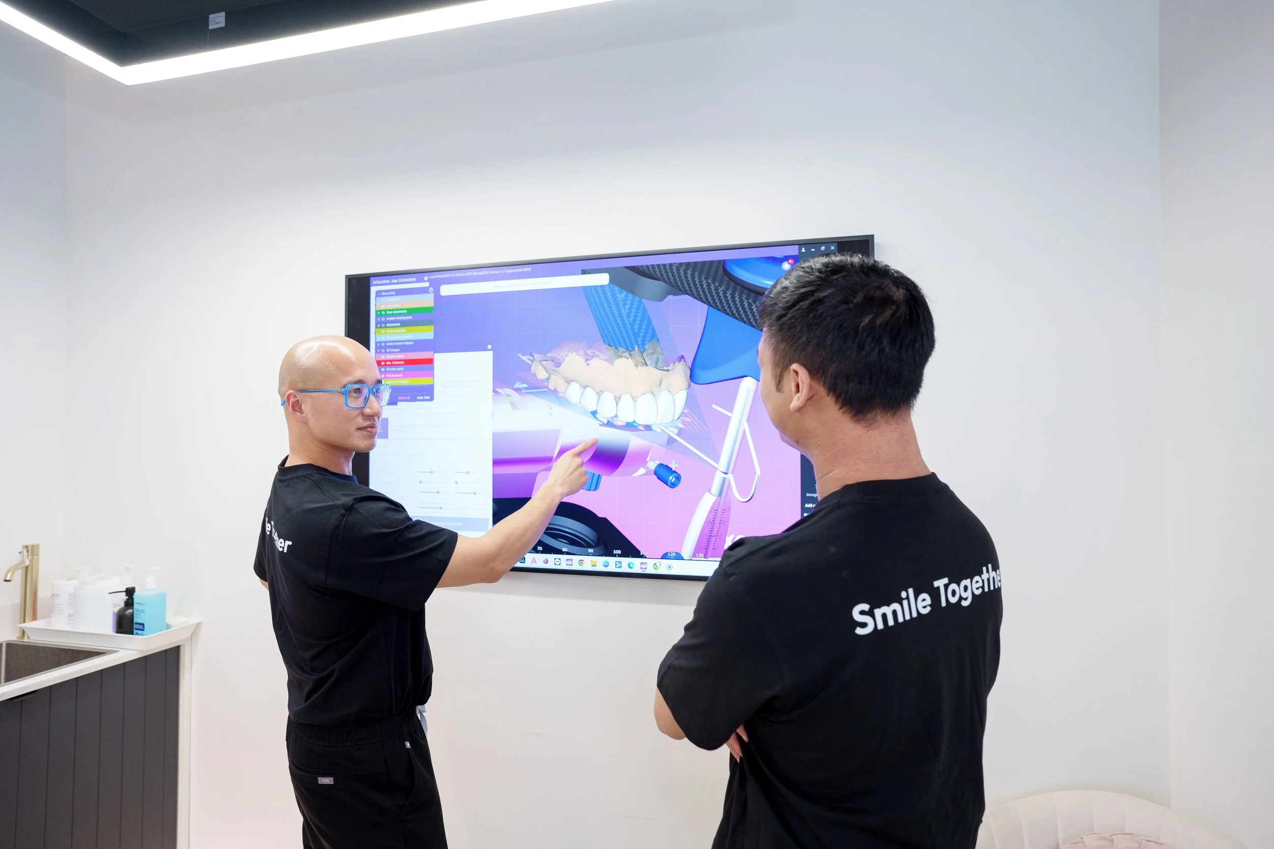 Two individuals in black shirts with 'Smile Together' written on the back, engaged in a discussion in front of a large digital screen displaying a 3D model of teeth, dental instruments, and colorful user interface elements in a clinical or educational setting.