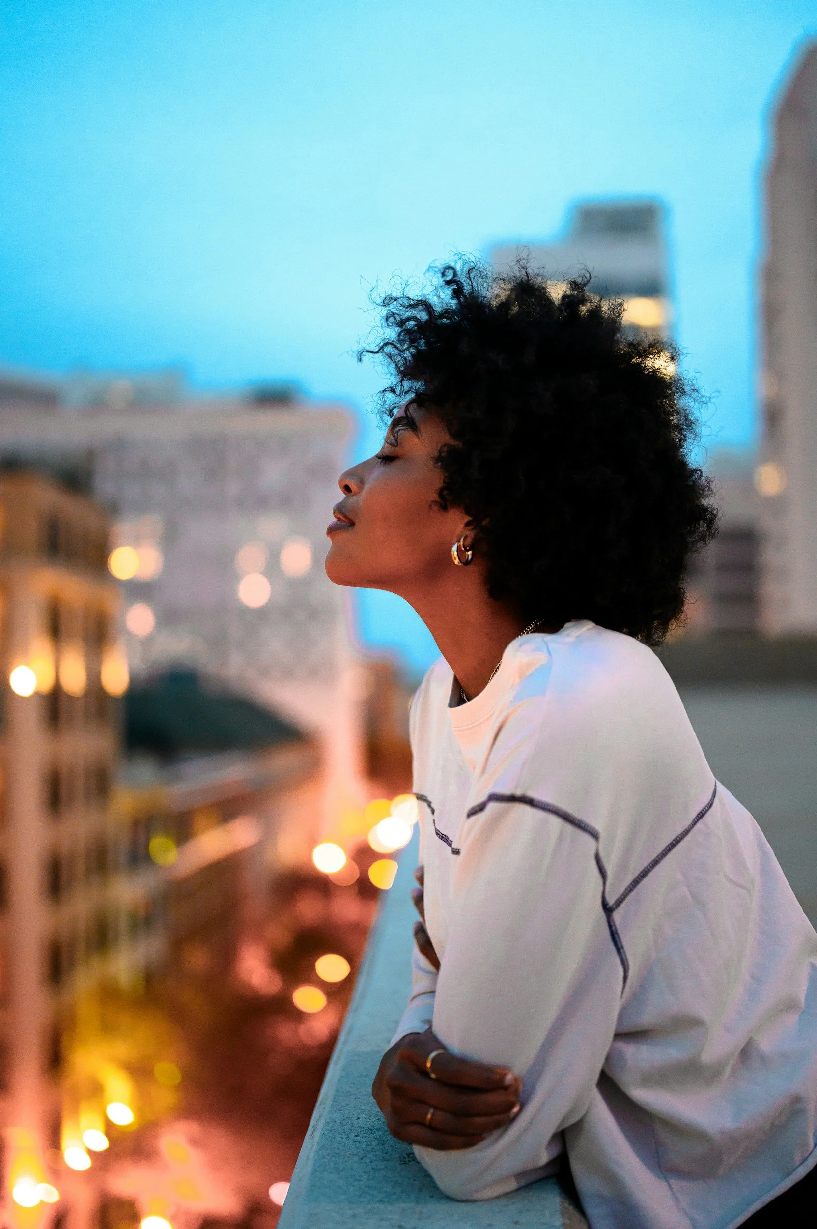 A woman with curly black hair wearing a white shirt with dark stripes, earrings, and rings, leaning on a railing with a cityscape and blurred lights in the background during dusk.