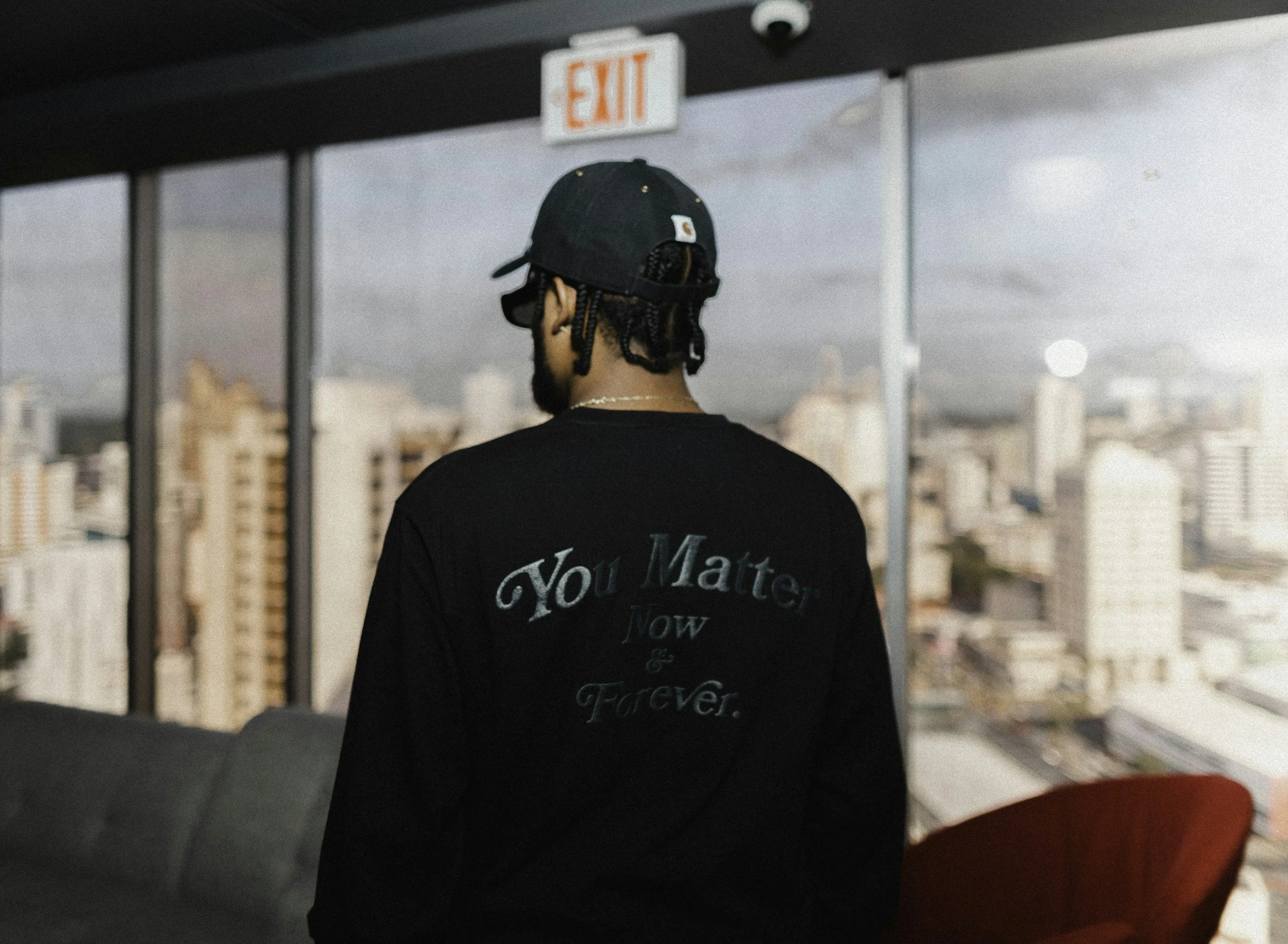 A person with braided hair wearing a black hoodie and a black cap with their back to the camera, looking out at a city skyline through large windows.