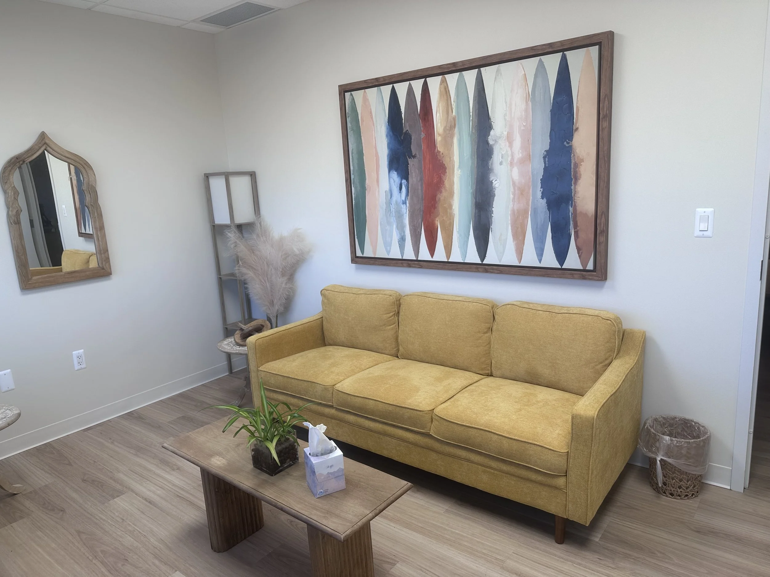 Living room with a mustard yellow sofa, wooden coffee table with a plant and tissue box, large colorful abstract painting on the wall, wooden framed mirror, tall frame holding dried pampas grass, small round side table, and a wicker wastebasket, with hardwood flooring.