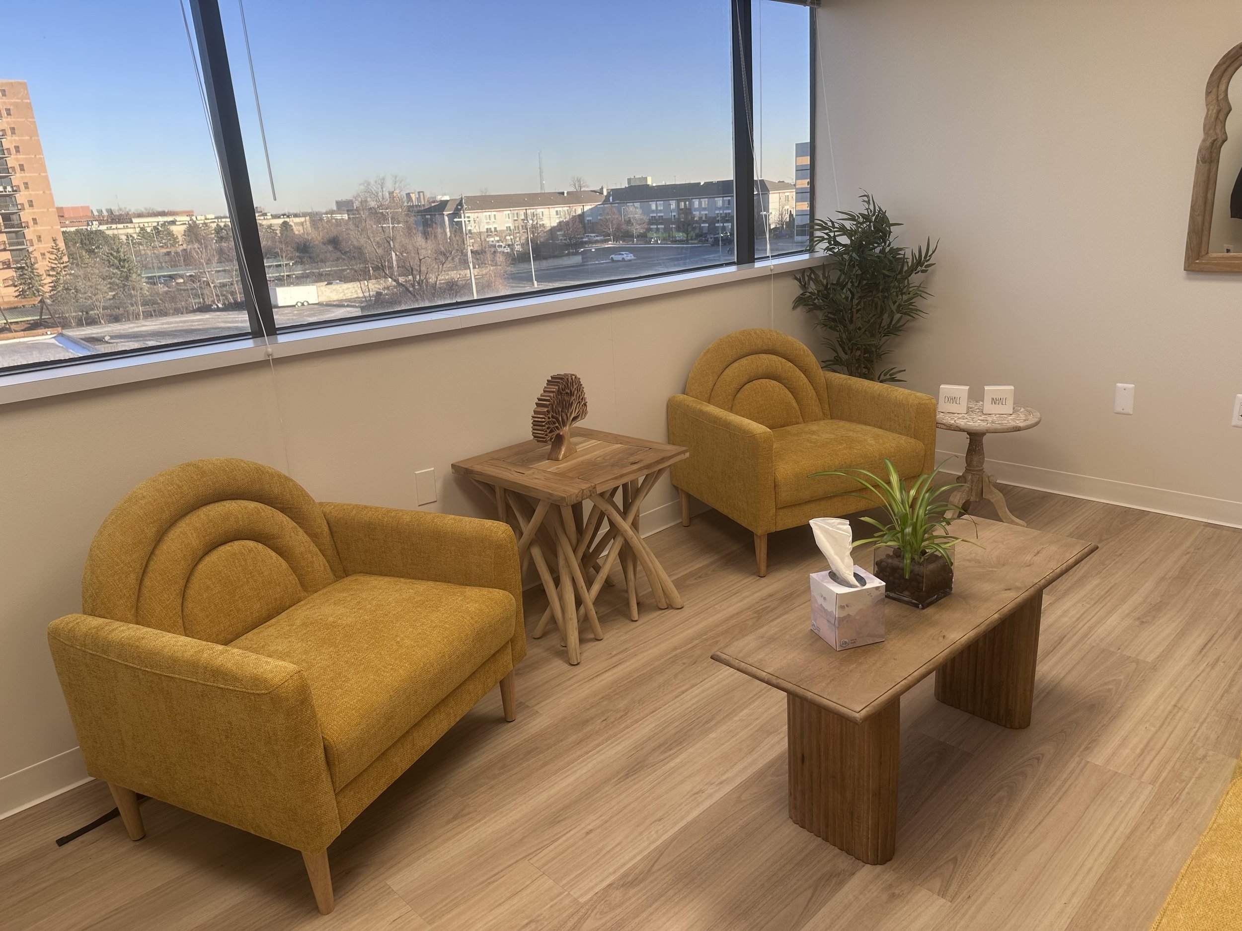 A cozy waiting room with two mustard yellow armchairs, a wooden coffee table with a potted plant and a box of tissues, a smaller wooden side table with decorative items, and a large window showing a cityscape with buildings and trees.