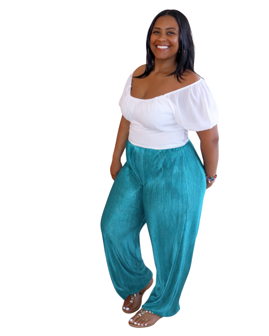 A smiling woman with dark hair, wearing a white peasant blouse and teal wide-leg pants, standing with one hand in her pocket.