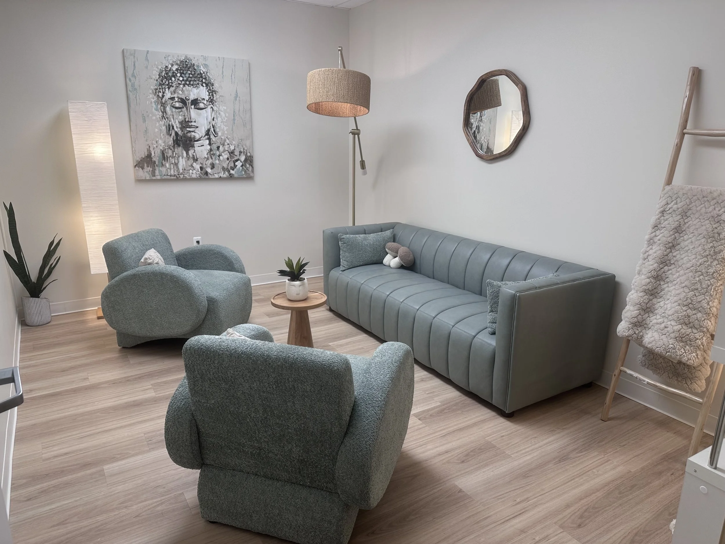 A living room with a light blue sofa, two matching armchairs, a small wooden coffee table with a plant, a tall lamp, a large Buddha artwork, a mirror, and a ladder with a blanket draped over it.