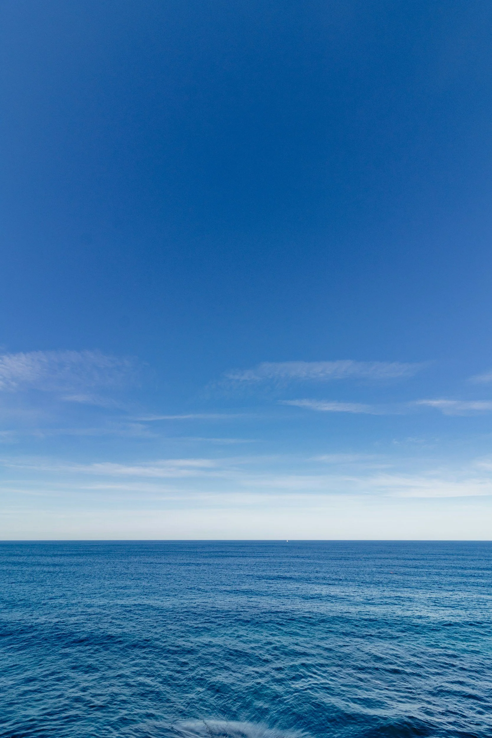 A clear blue sky with some white clouds over a vast ocean with gentle waves.