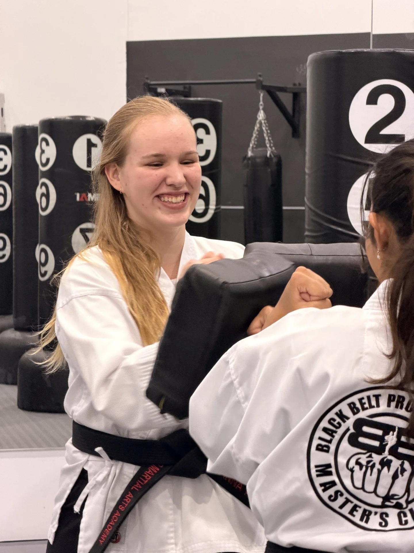 Close-up of a smiling woman with curly hair wearing glasses, hoop earrings, and layered necklaces, in an indoor setting.